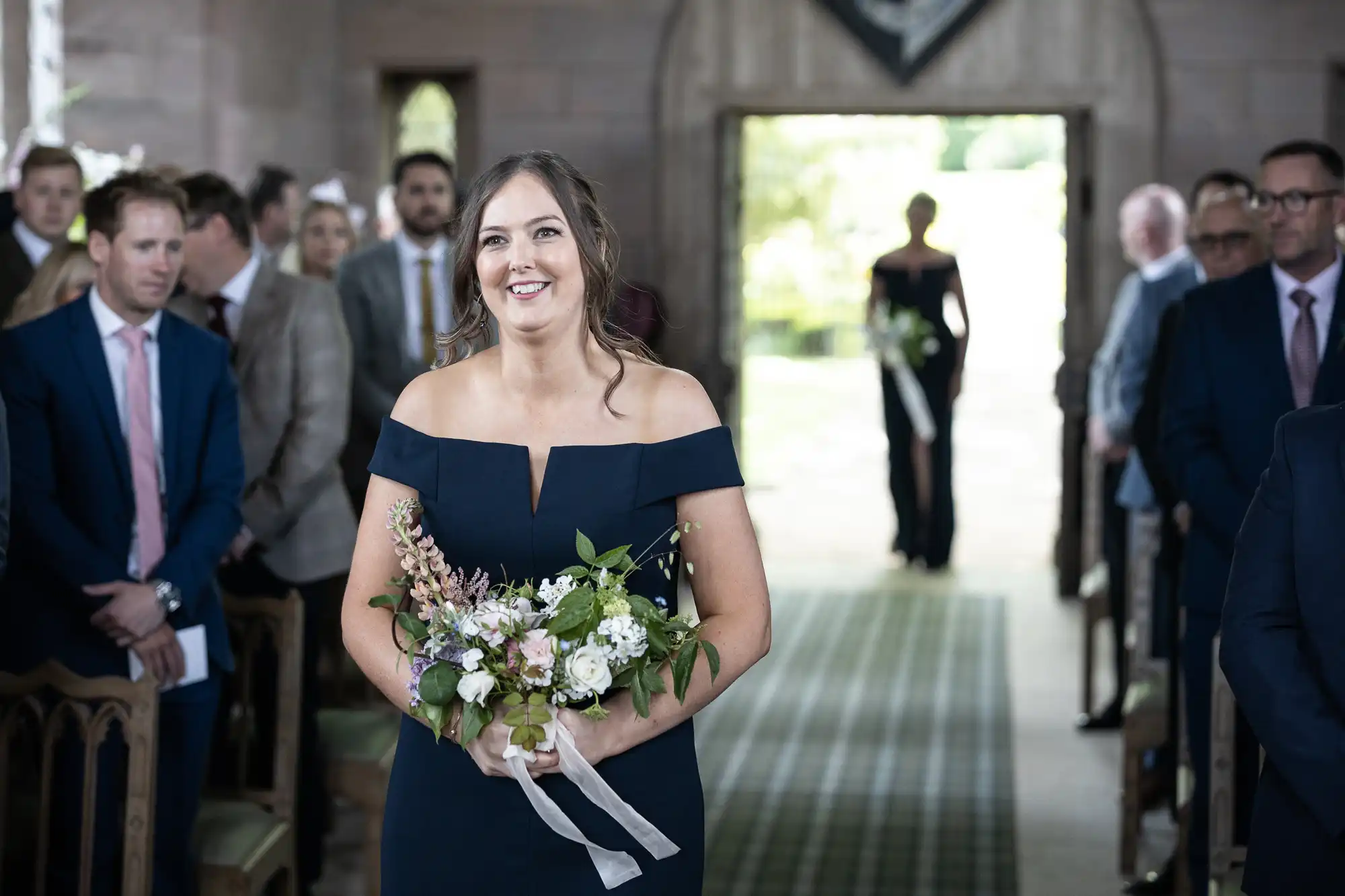 A bridesmaid in a navy dress holds a bouquet, smiling as she walks down the aisle in a church, guests watching.