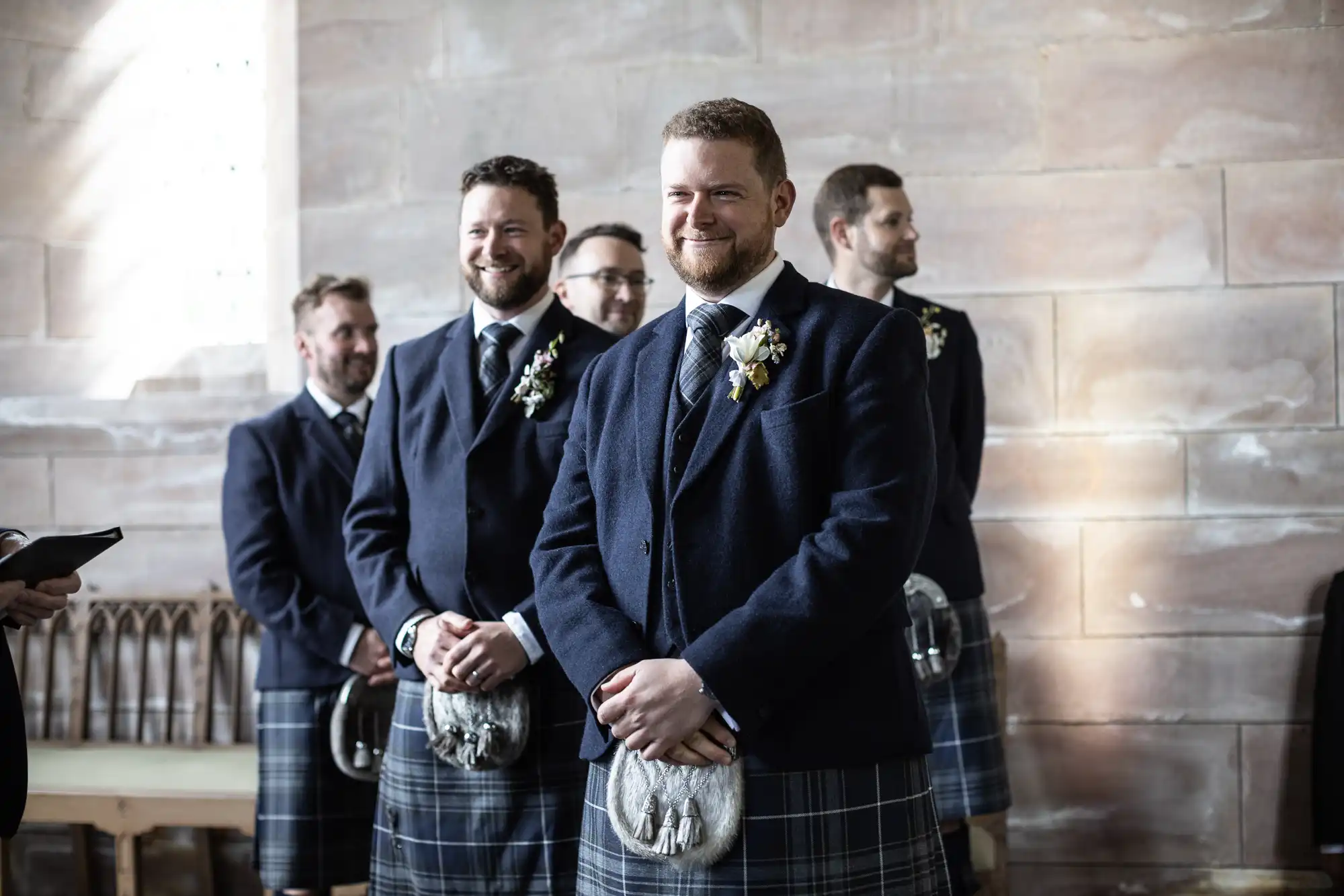 Groom smiling in kilt with groomsmen in background at wedding ceremony.