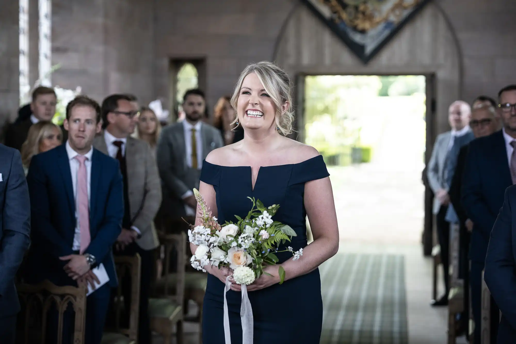 A smiling woman in a dark blue dress holding a bouquet walks down the aisle of a church, with seated guests watching.
