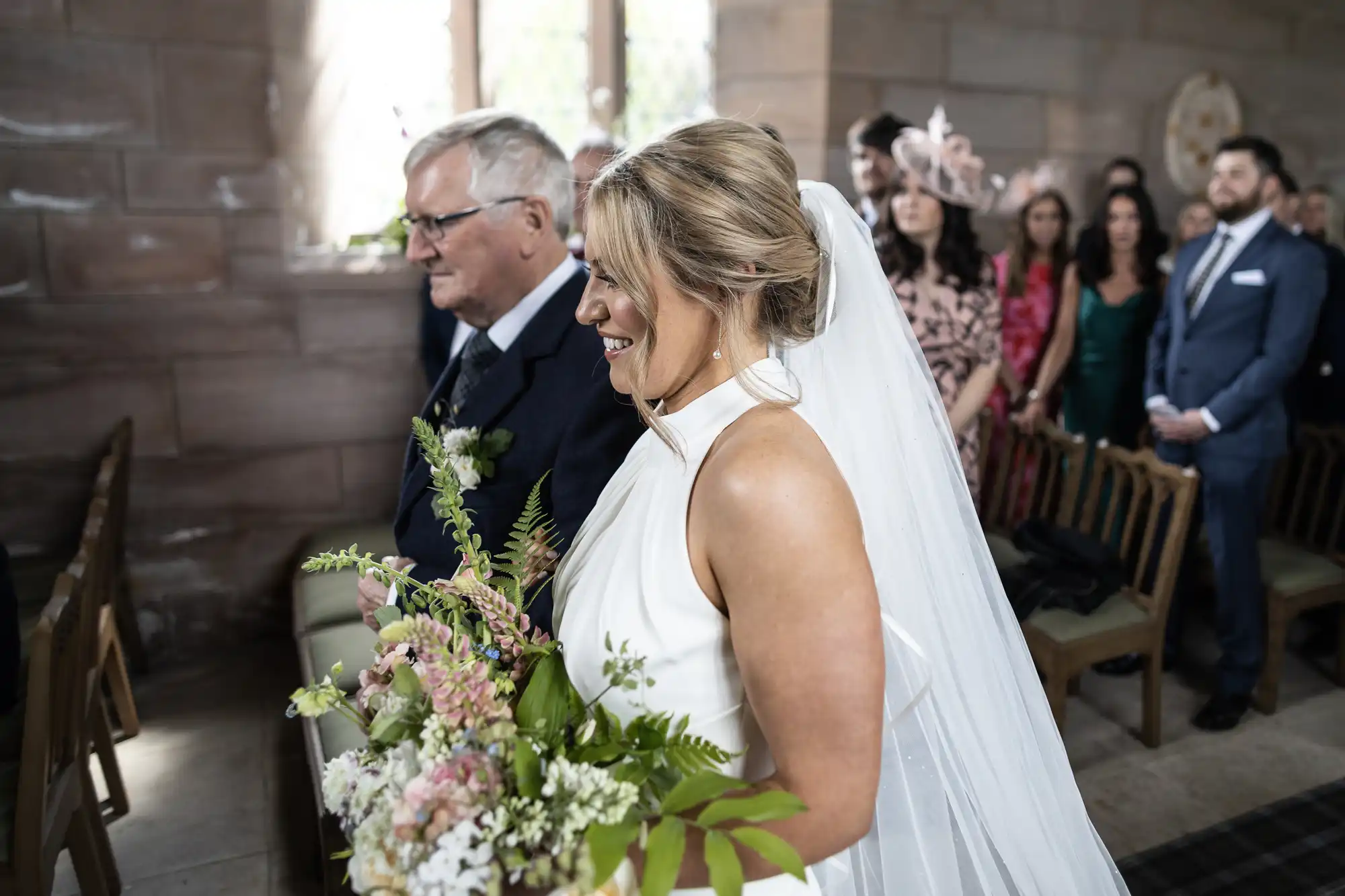 A bride in a white dress holding a bouquet walks down the aisle in a church, accompanied by an older man, with guests watching.