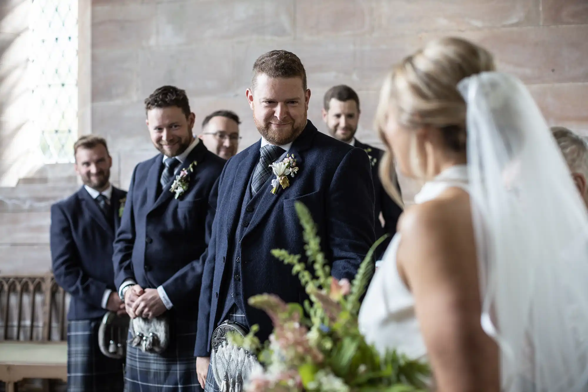 A groom in a blue suit and kilt smiles at the bride during a wedding ceremony in a church, with guests in kilts watching in the background.