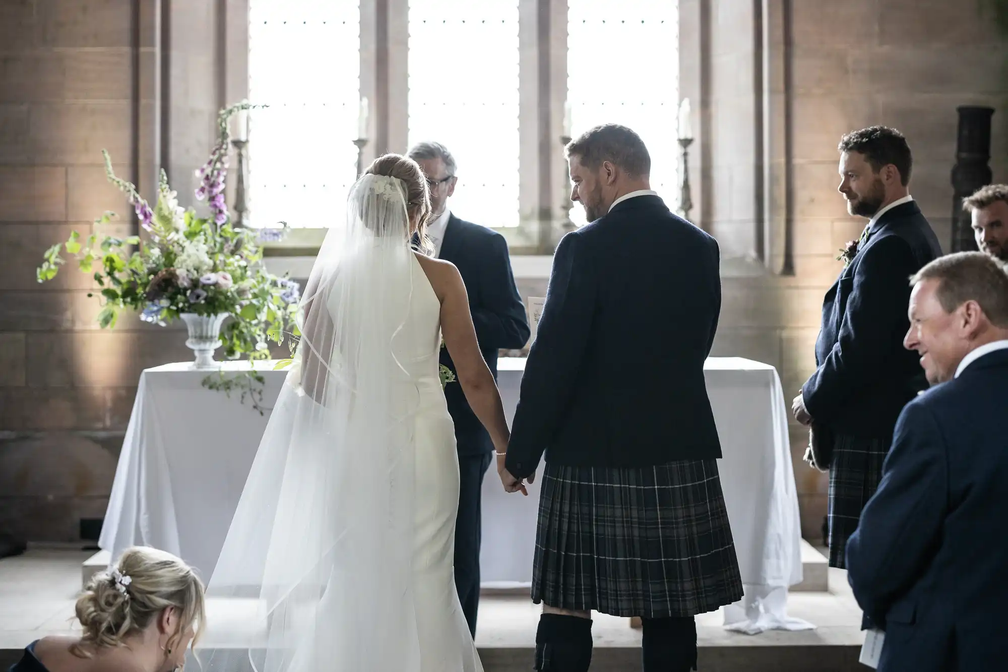 A bride and groom holding hands at the altar during a wedding ceremony, with the groom wearing a kilt.