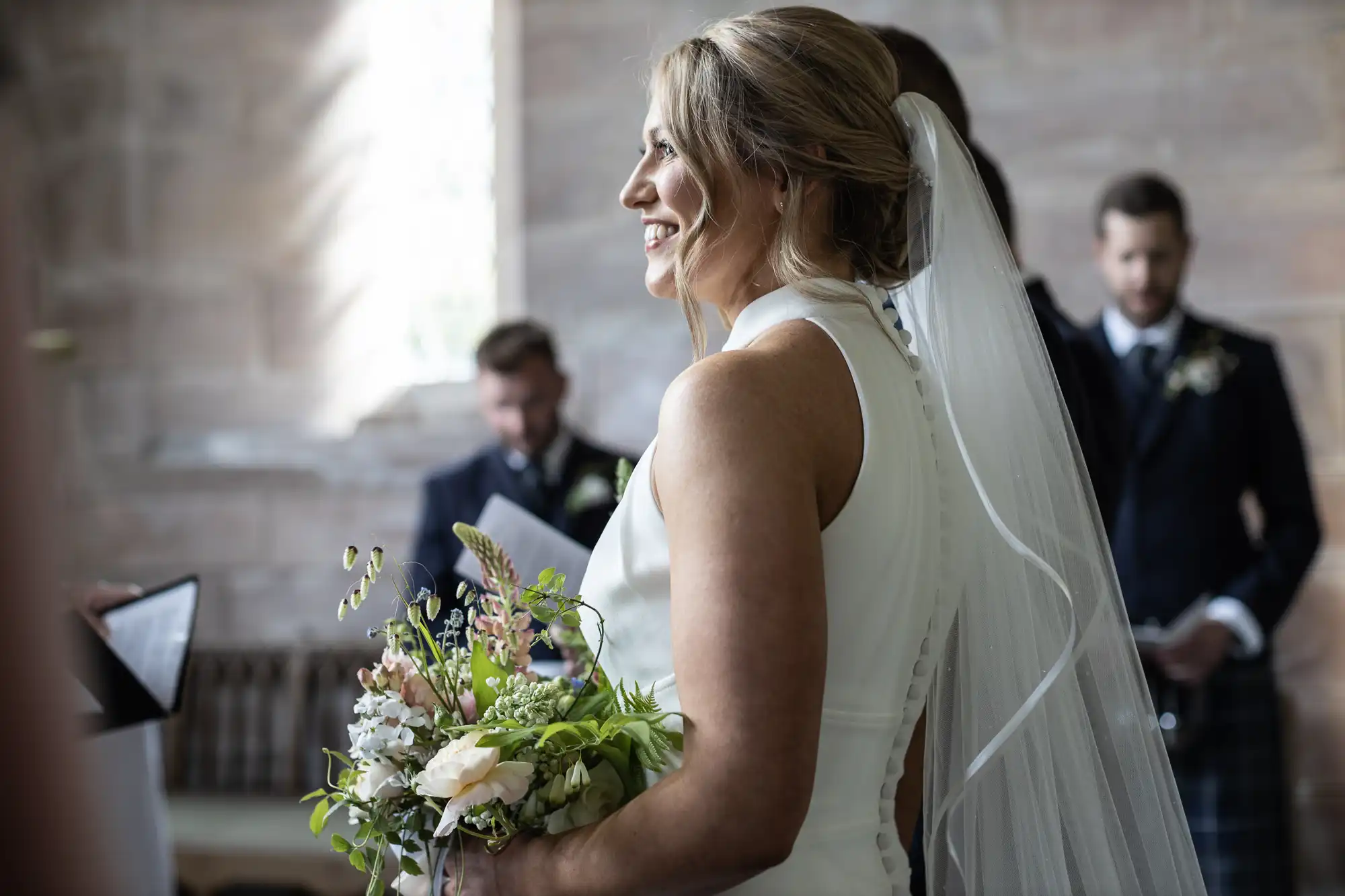 A bride holding a bouquet smiles during a wedding ceremony, with the groom looking on in the background in a church.
