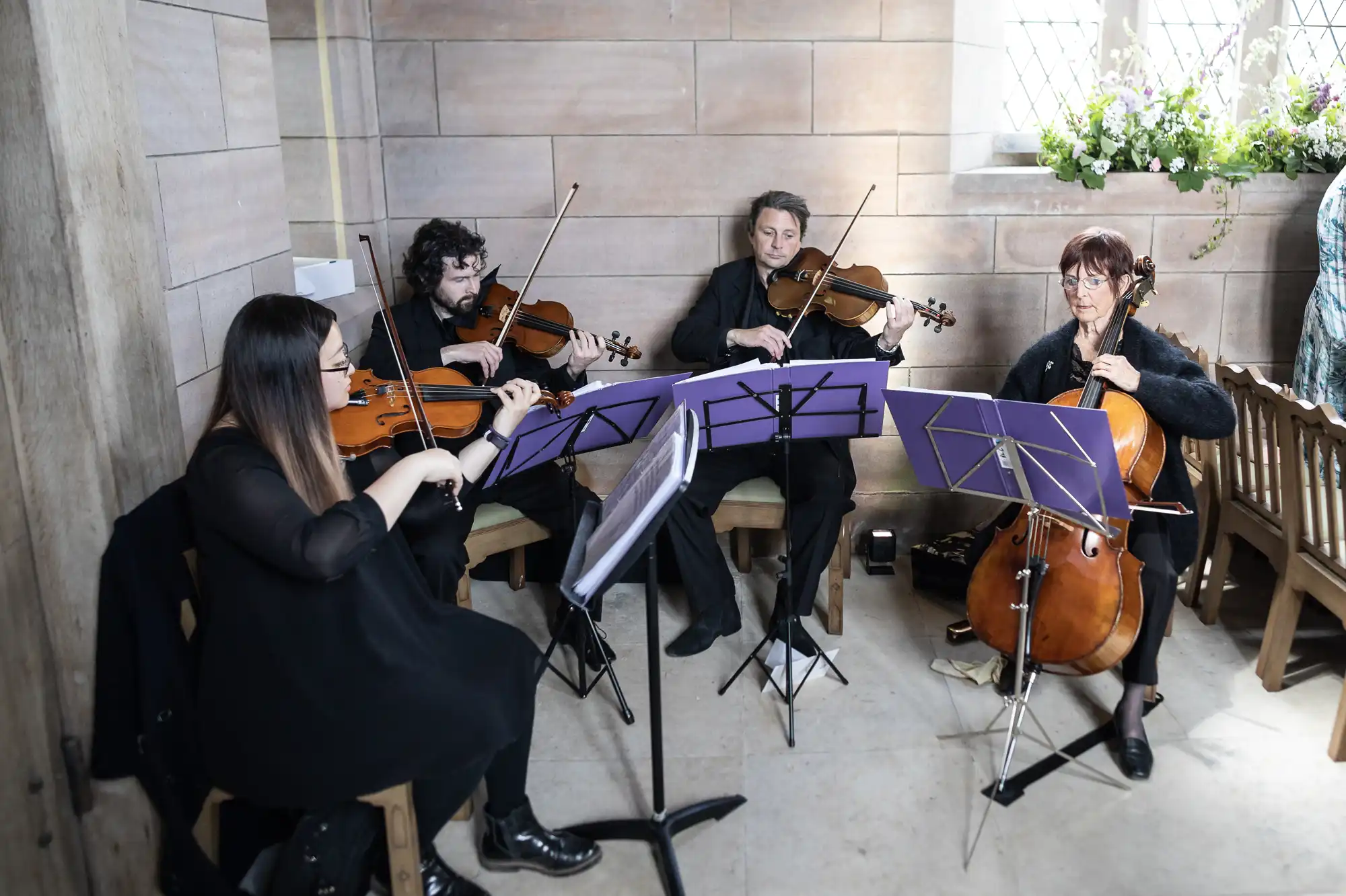 A string quartet with two violinists, a violist, and a cellist performing indoors, using sheet music on stands.
