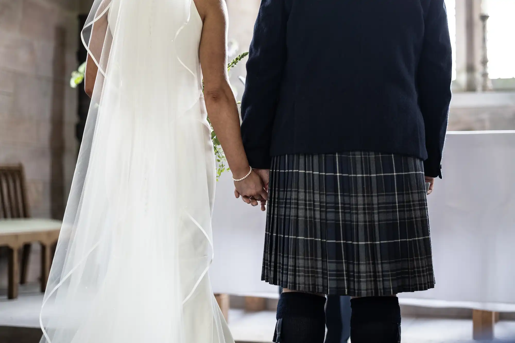 A bride in a white gown and a groom in a kilt hold hands during a wedding ceremony in a church.