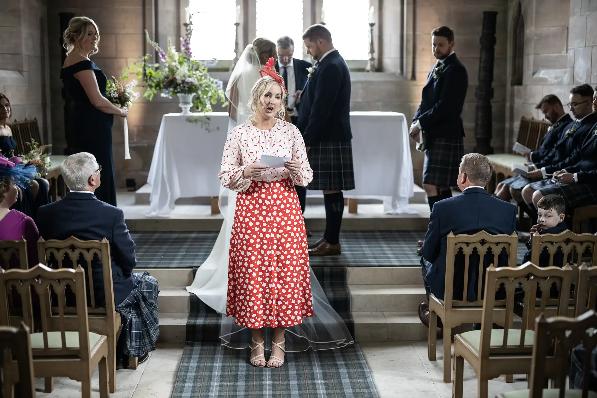 A woman in a red polka dot dress stands reading from a sheet of paper at a wedding ceremony in a stone hall, with guests in formal attire listening attentively.