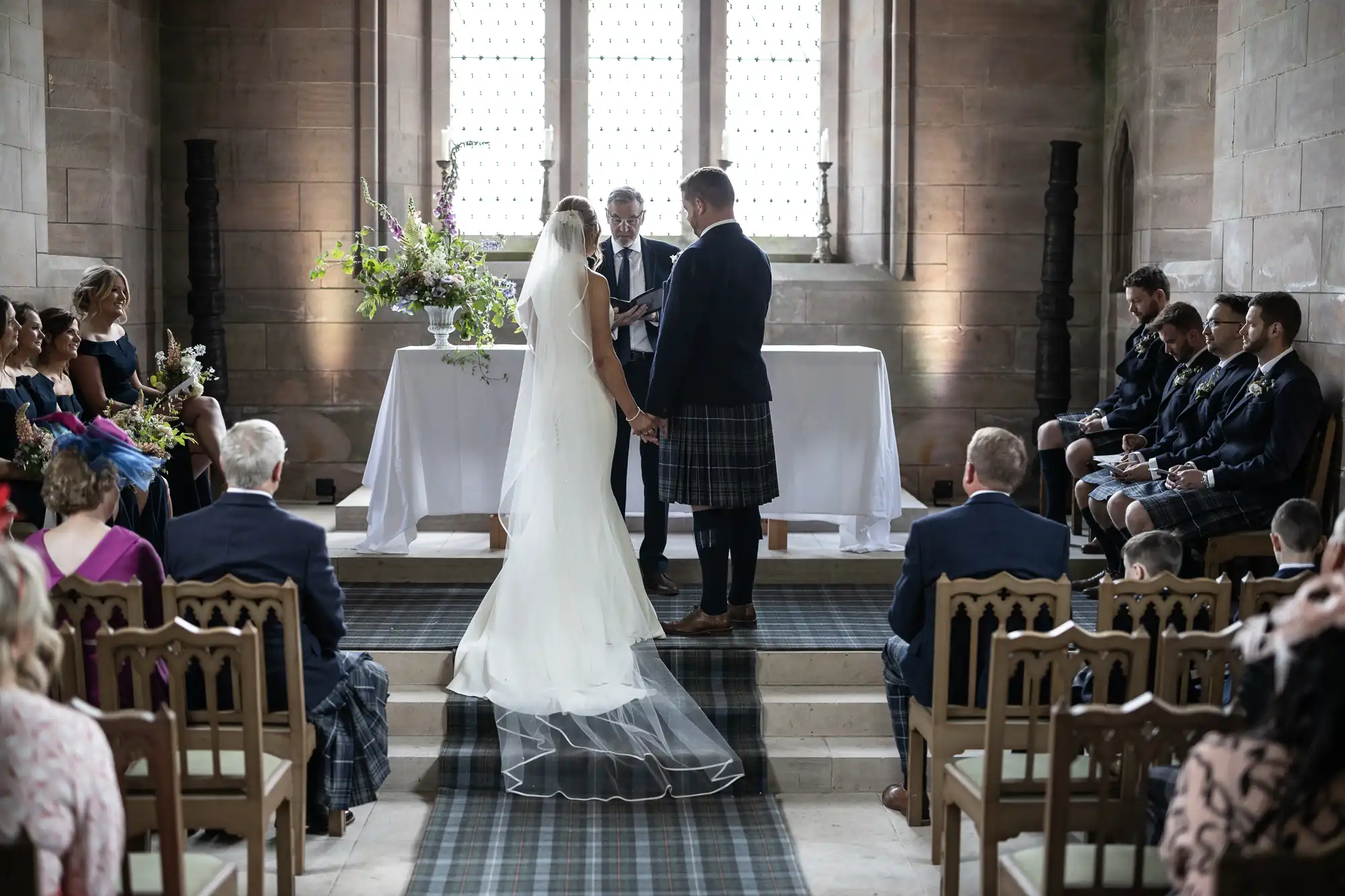 A bride and groom exchanging vows in a church, with guests in kilts observing from wooden pews.