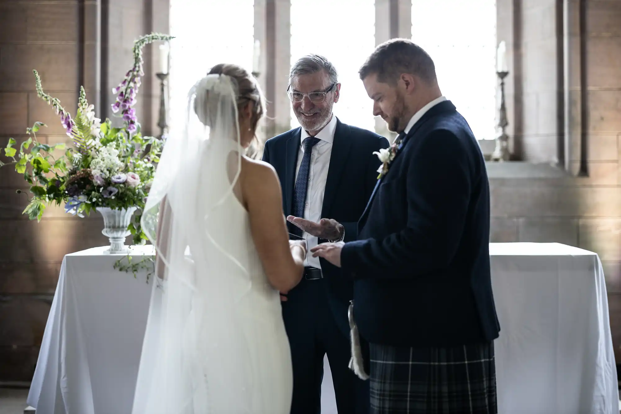 A bride and groom exchange vows at a wedding ceremony, overseen by an officiant in a suit, in a softly lit hall with floral decorations.