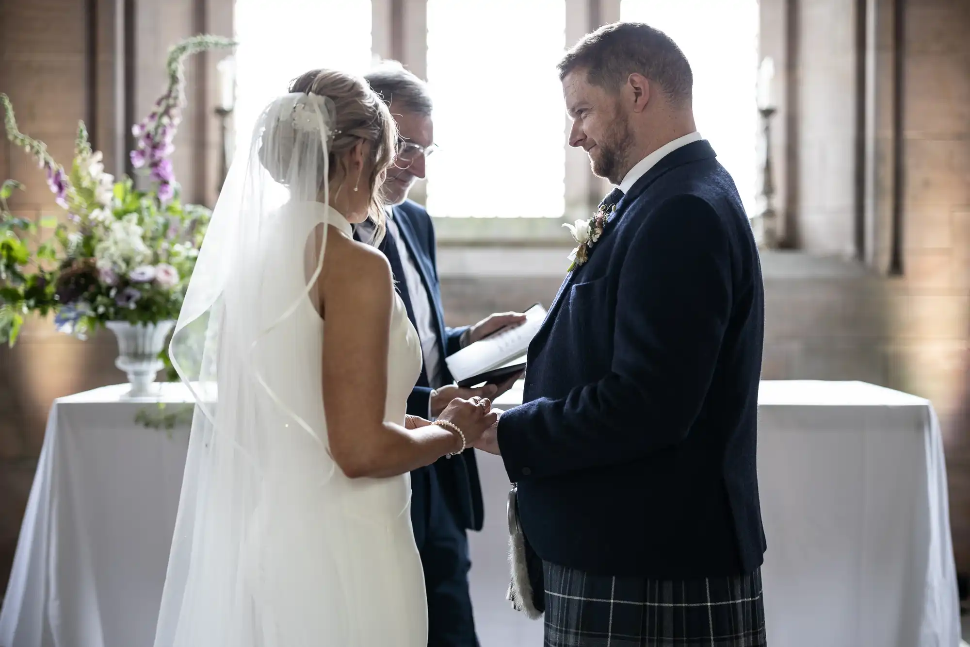A bride and groom exchanging rings during a wedding ceremony, officiated by a woman, inside a sunlit room.