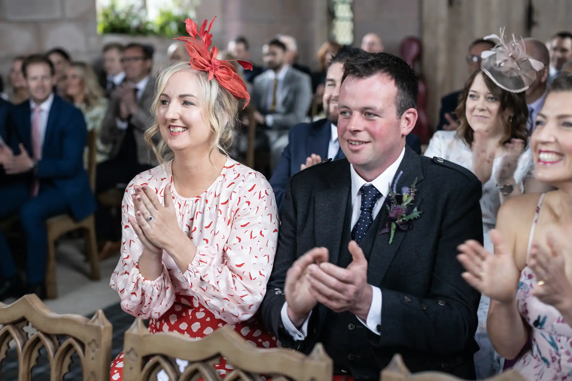 Guests in formal attire clapping at a wedding ceremony, with a woman in a red and white dress and a red hat, and a man in a suit with a flower boutonniere.