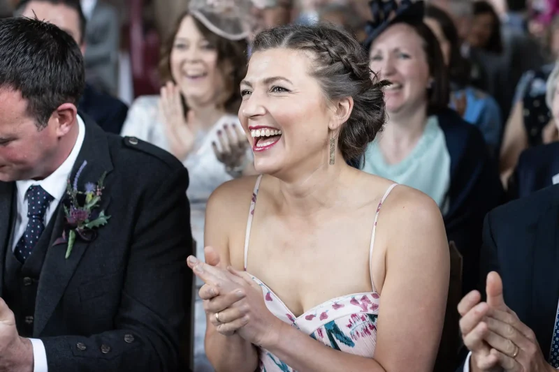 A joyful woman in a floral dress clapping at a wedding ceremony, surrounded by other guests.