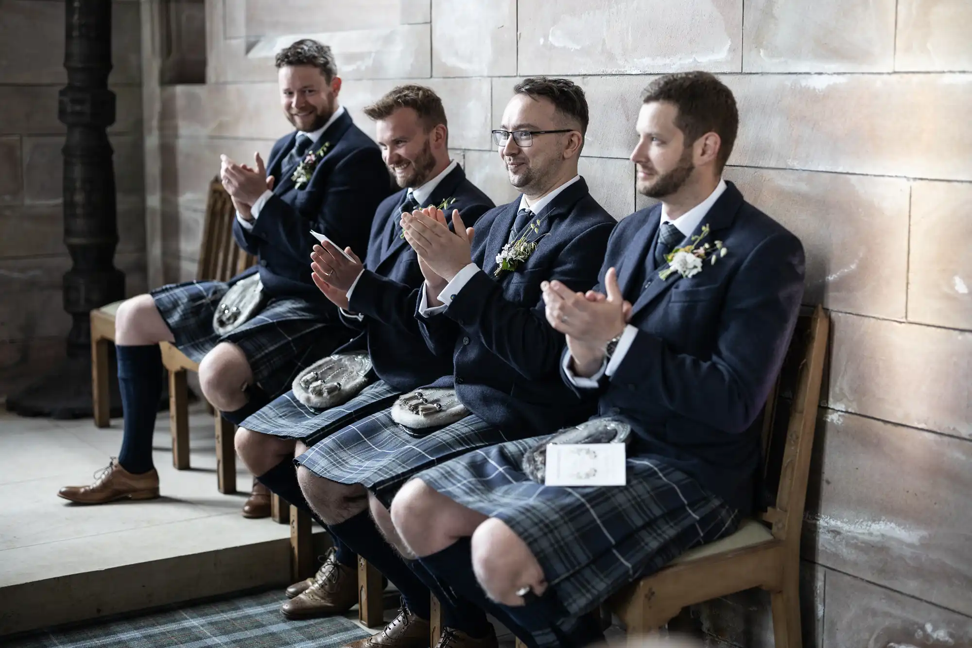 Four men in traditional kilts and blazers clapping while seated on a stone bench indoors.