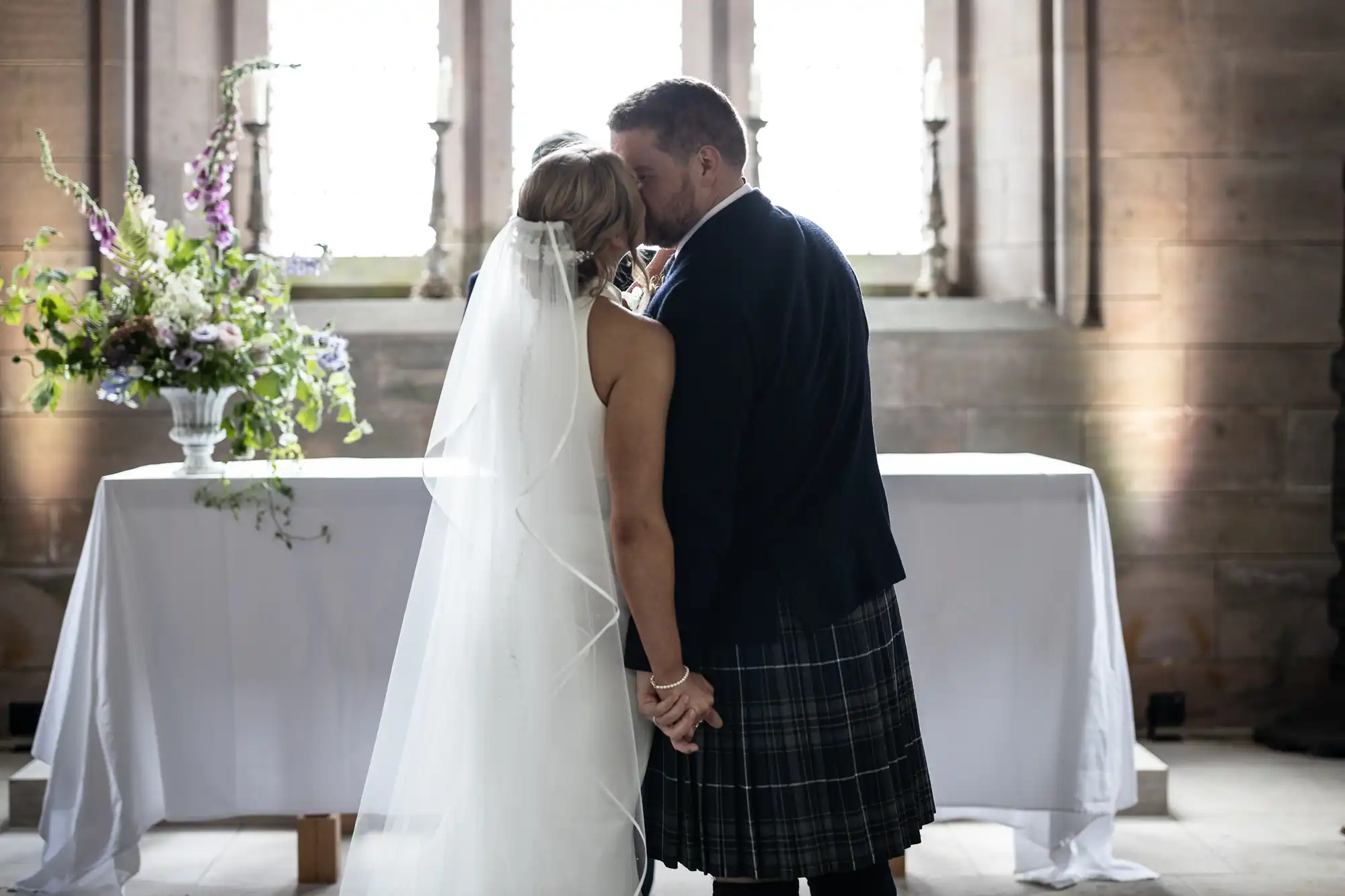 Bride and groom kissing in a church, groom in a kilt, with a table adorned with a floral arrangement in the background.