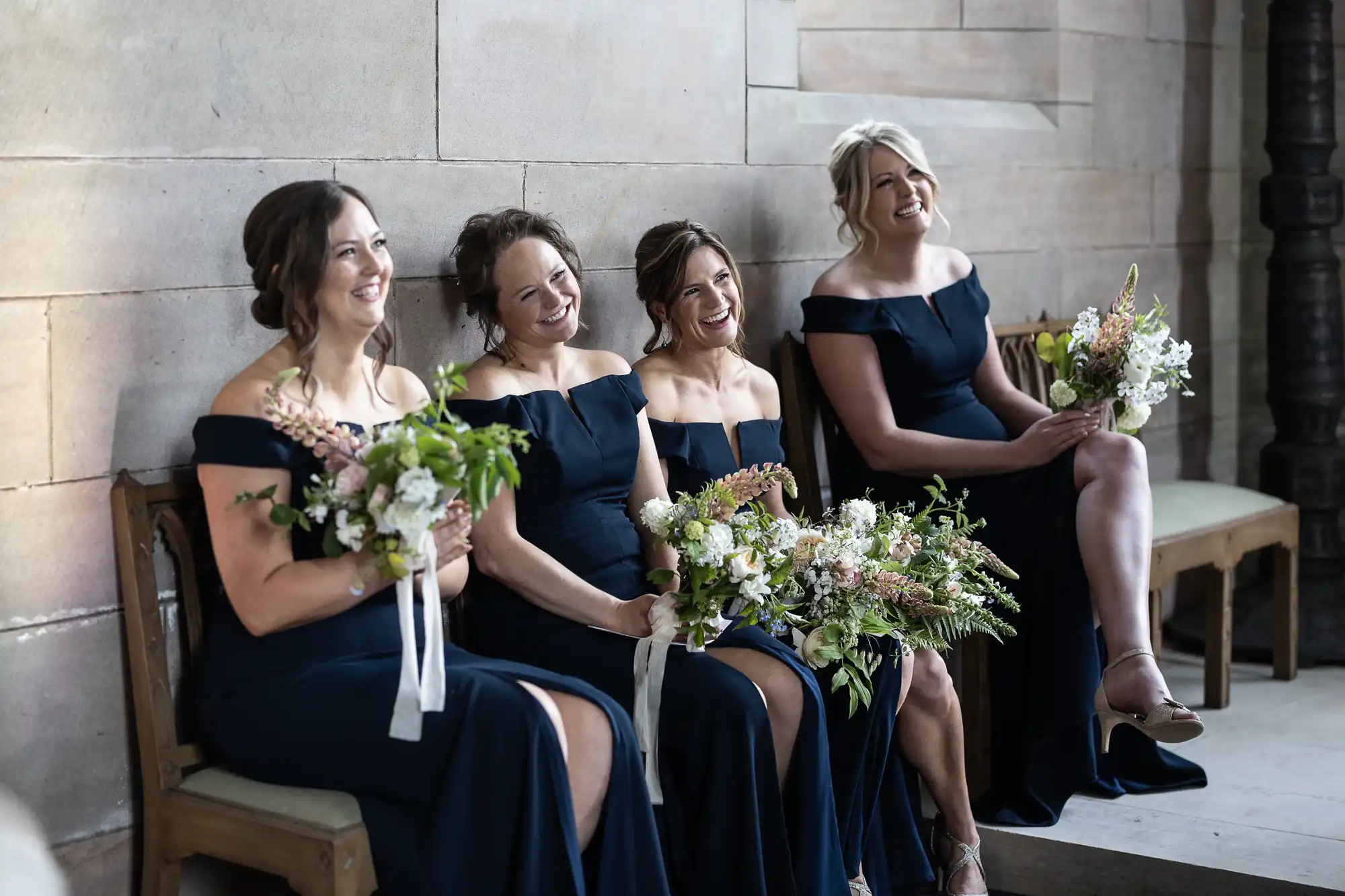 Four women in navy blue dresses sitting side by side, smiling and holding bouquets, in a grand stone hall.