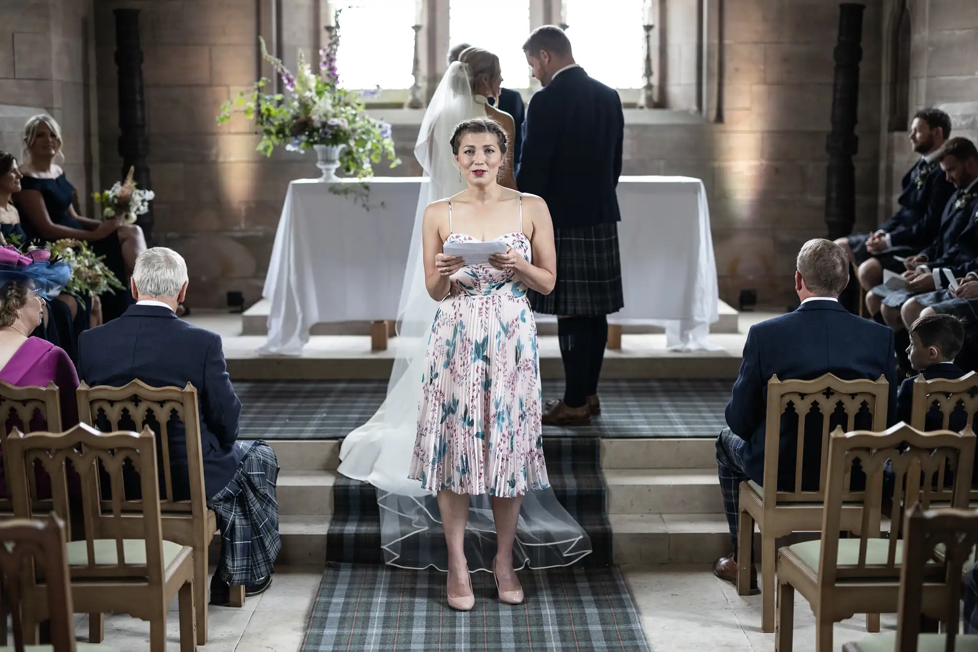 A woman reads from a card at a wedding ceremony in a church, with guests seated around and a couple at the altar in the background.