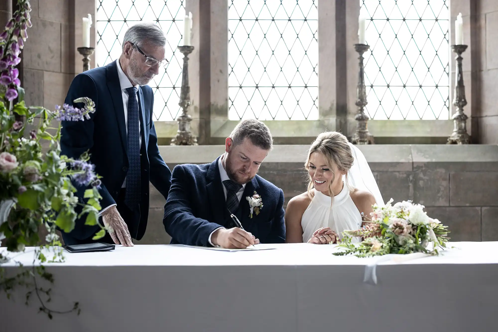 A bride and groom smiling as the groom signs a document at a wedding ceremony table, with an officiant standing next to them in a historic building with stained glass windows.