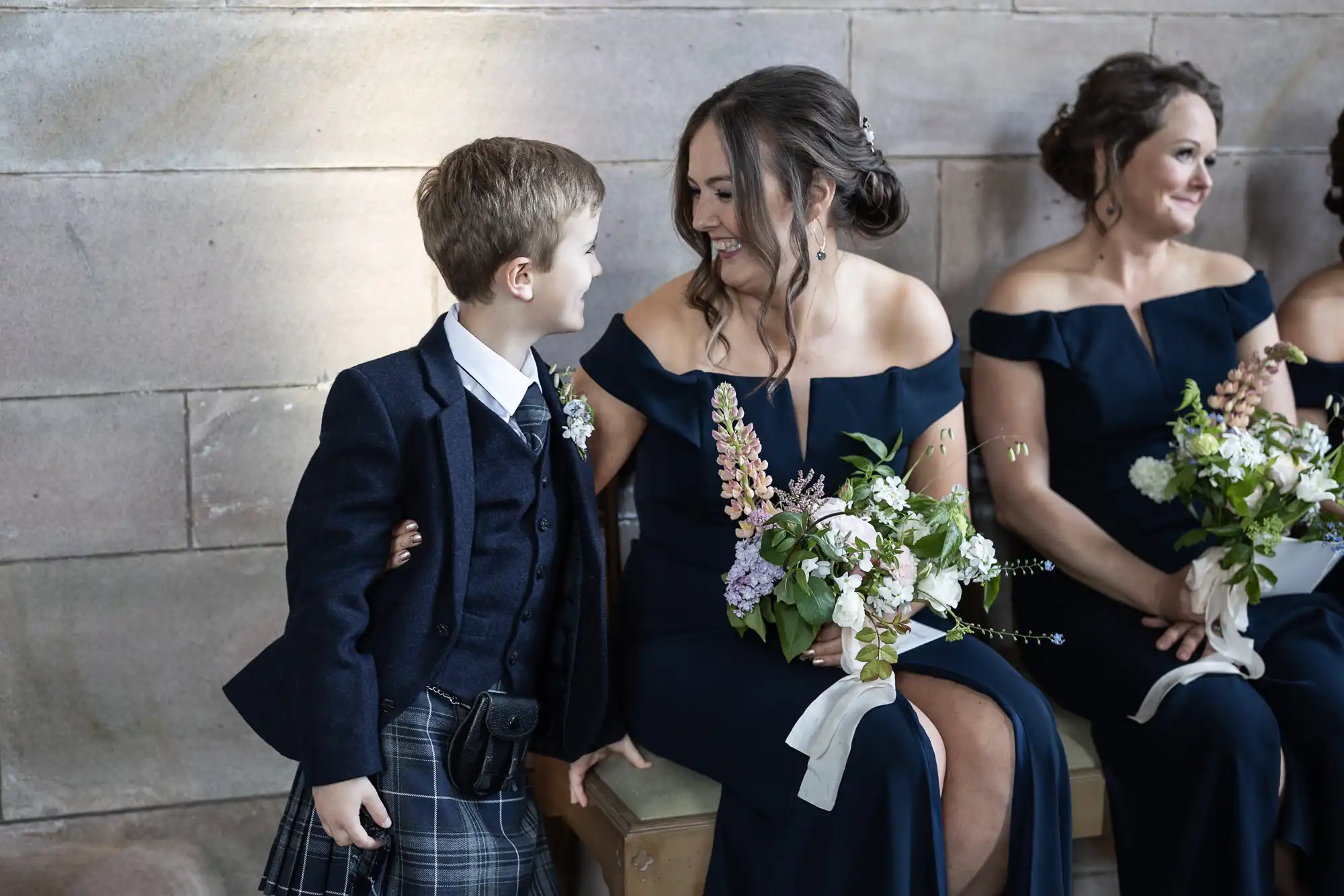 A young boy in a kilt smiles at a woman in a navy dress holding flowers, sitting beside another woman with flowers at a formal event.