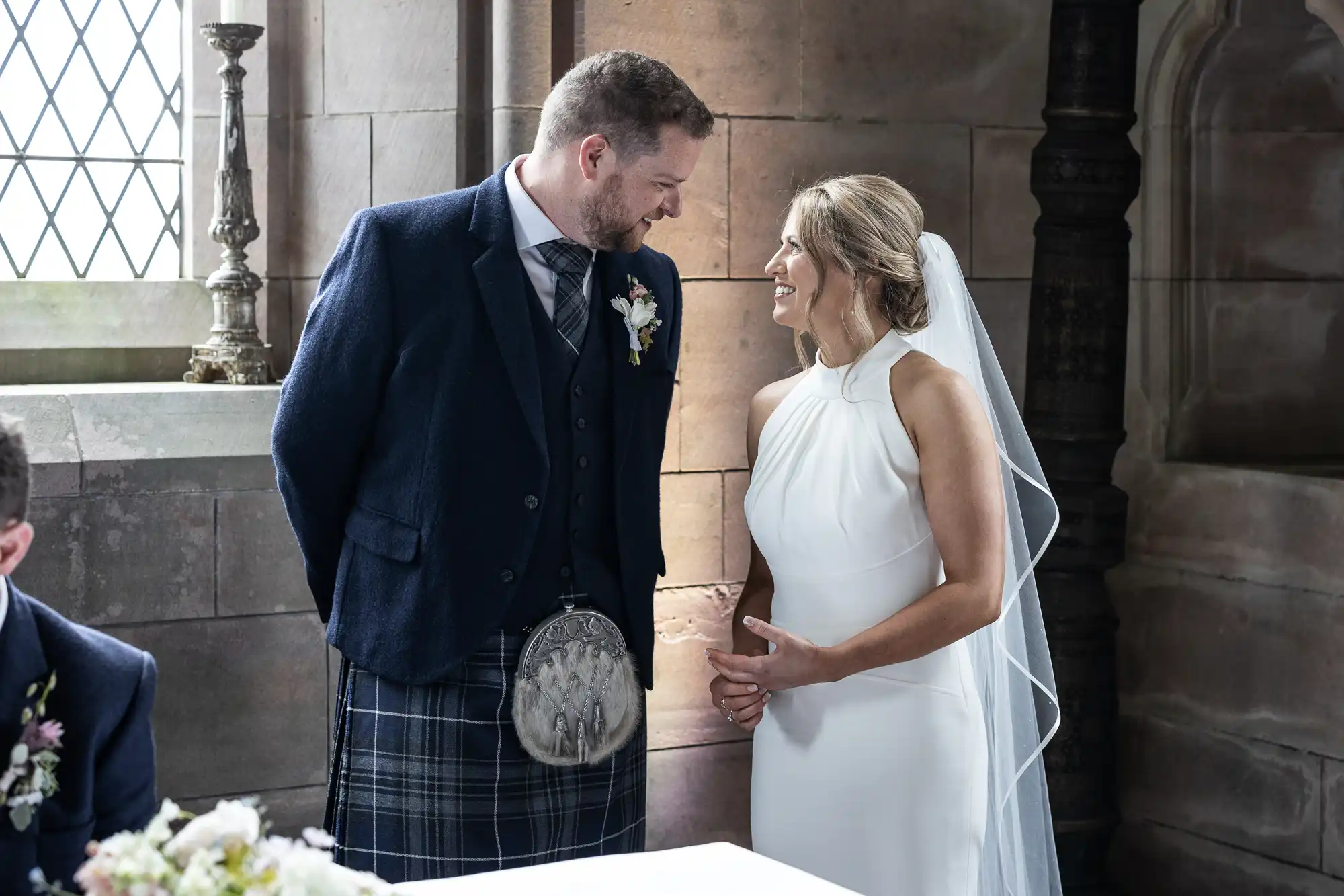 A bride and groom smiling at each other during their wedding ceremony inside a church, with the groom wearing traditional Scottish tartan attire.