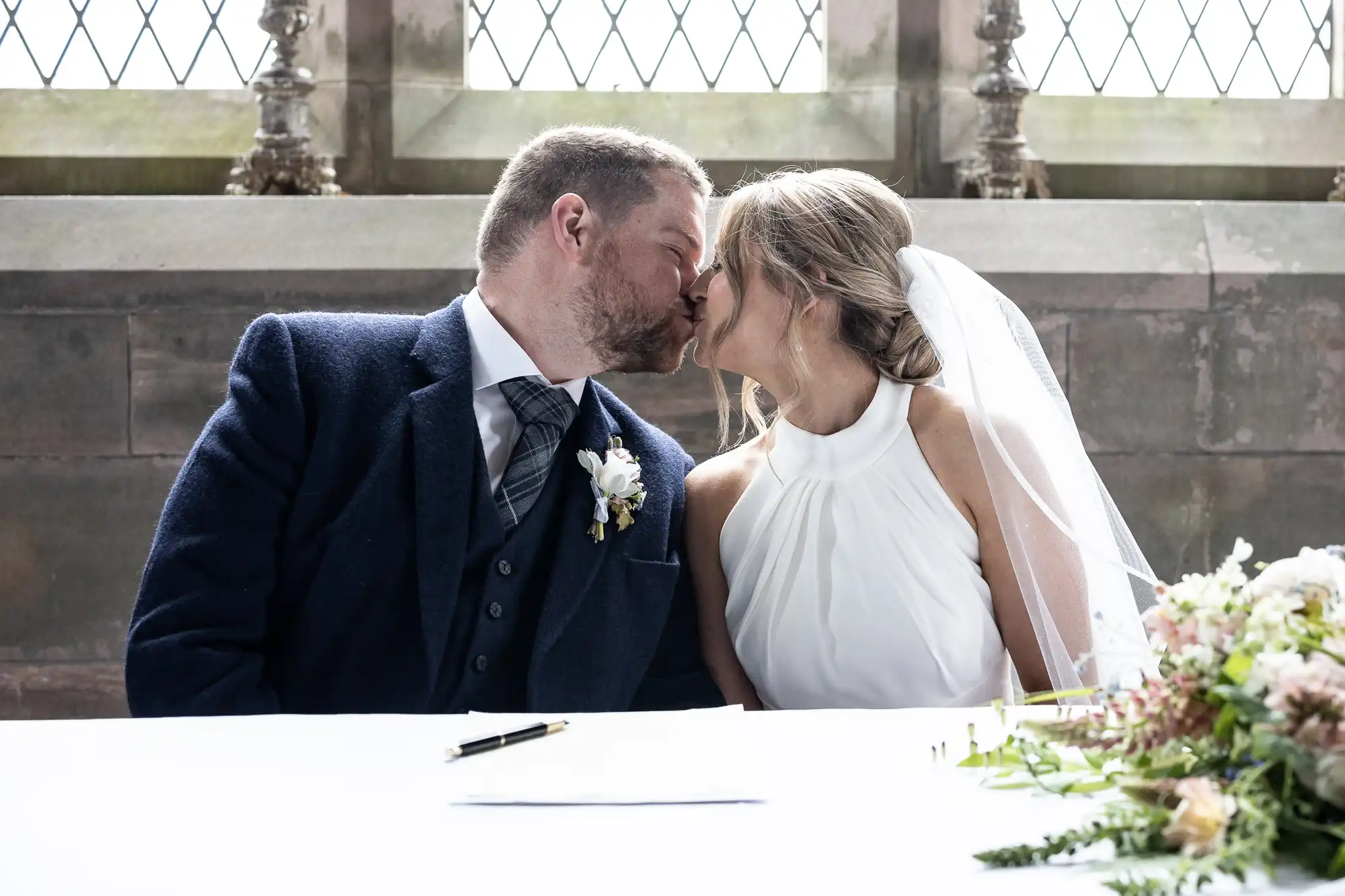 Bride and groom kissing at the signing table in a church, with natural light streaming through windows and flowers beside them.