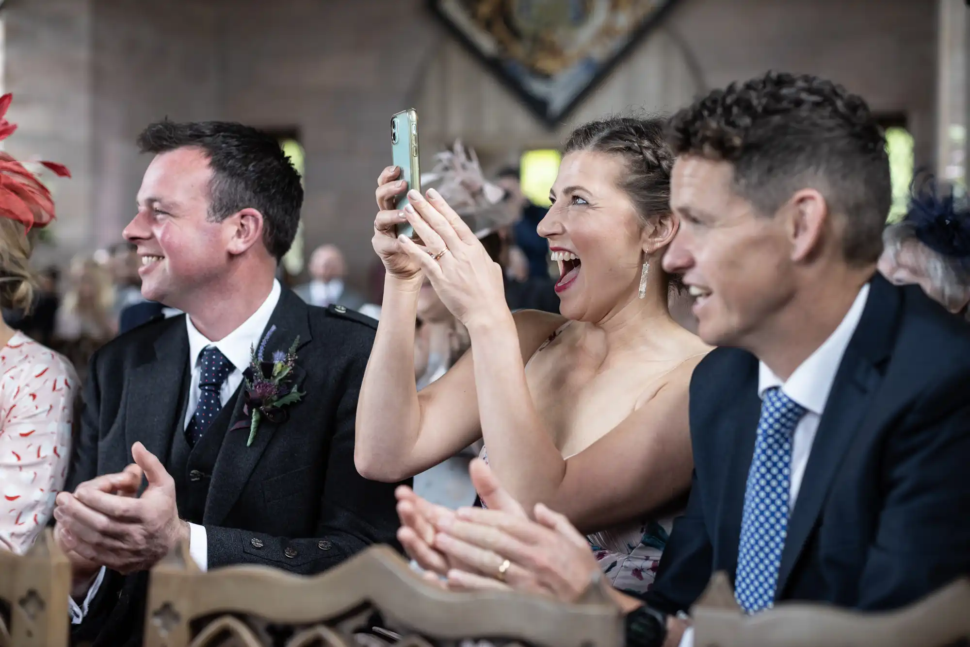 A woman joyfully recording a wedding ceremony on her smartphone, flanked by two smiling men in suits and guests in formal attire.