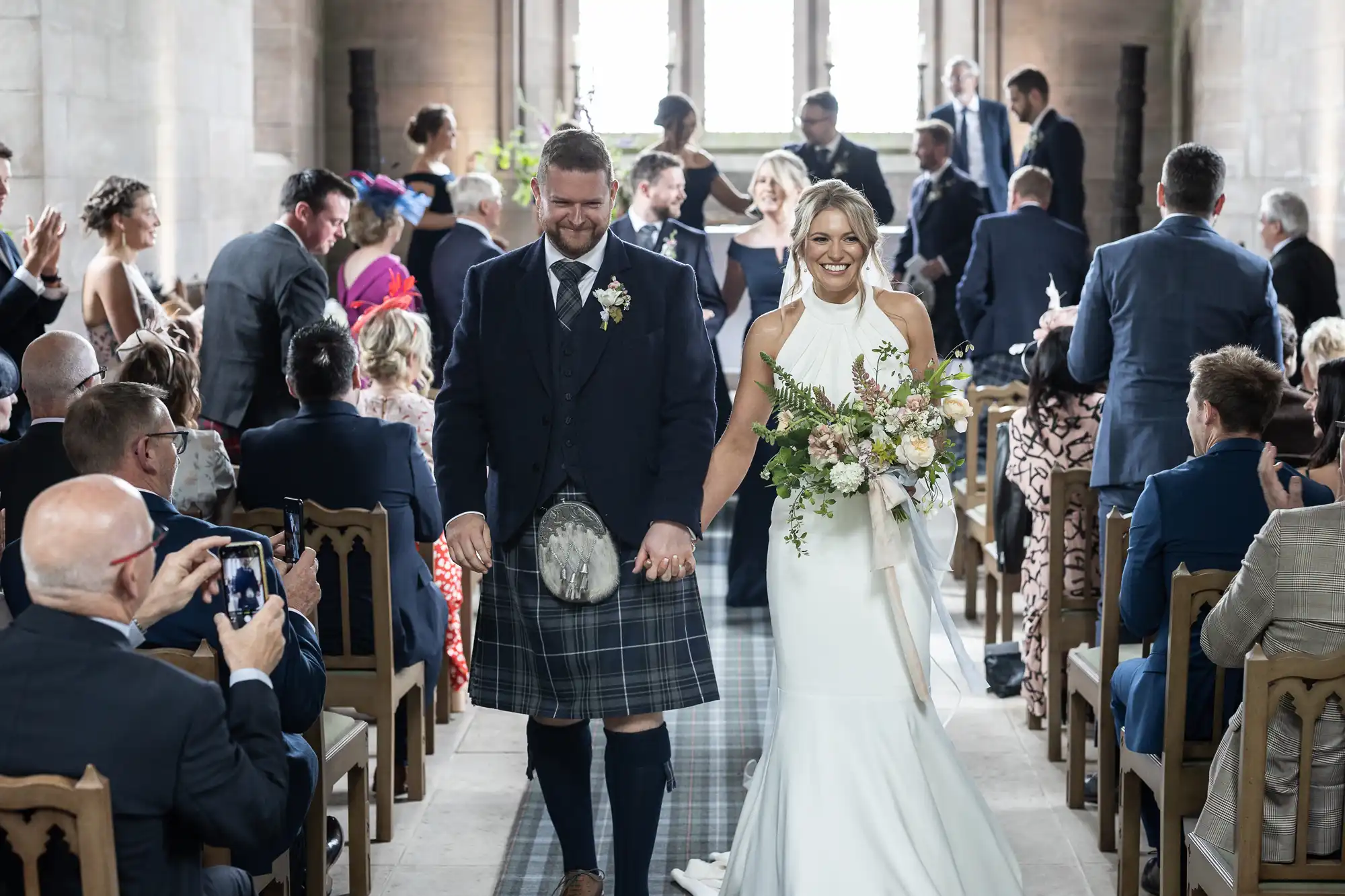 A bride and groom walk down the aisle, smiling, as guests applaud; the groom wears a kilt and the bride carries a bouquet.