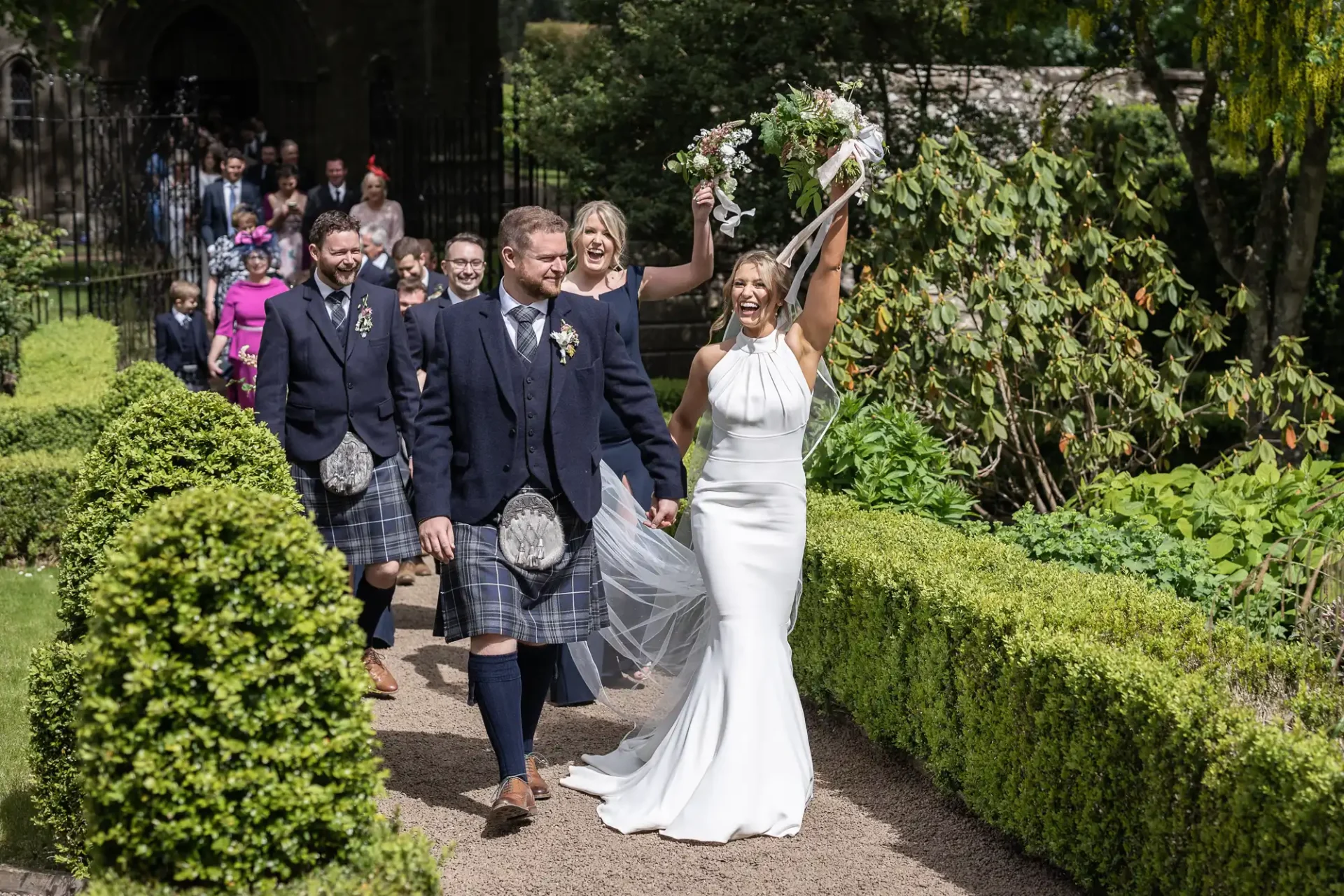 A joyful bride and groom walk hand in hand, leading guests down a garden path after their ceremony.