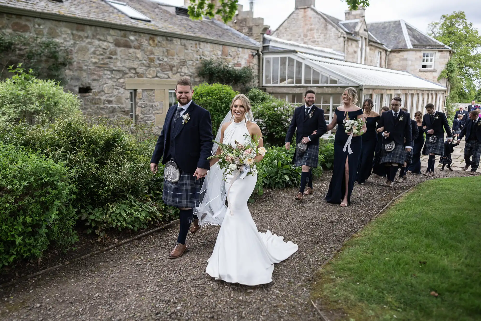 A newlywed couple leads a group while walking along a garden path, the groom wearing a kilt and the bride in a white gown.