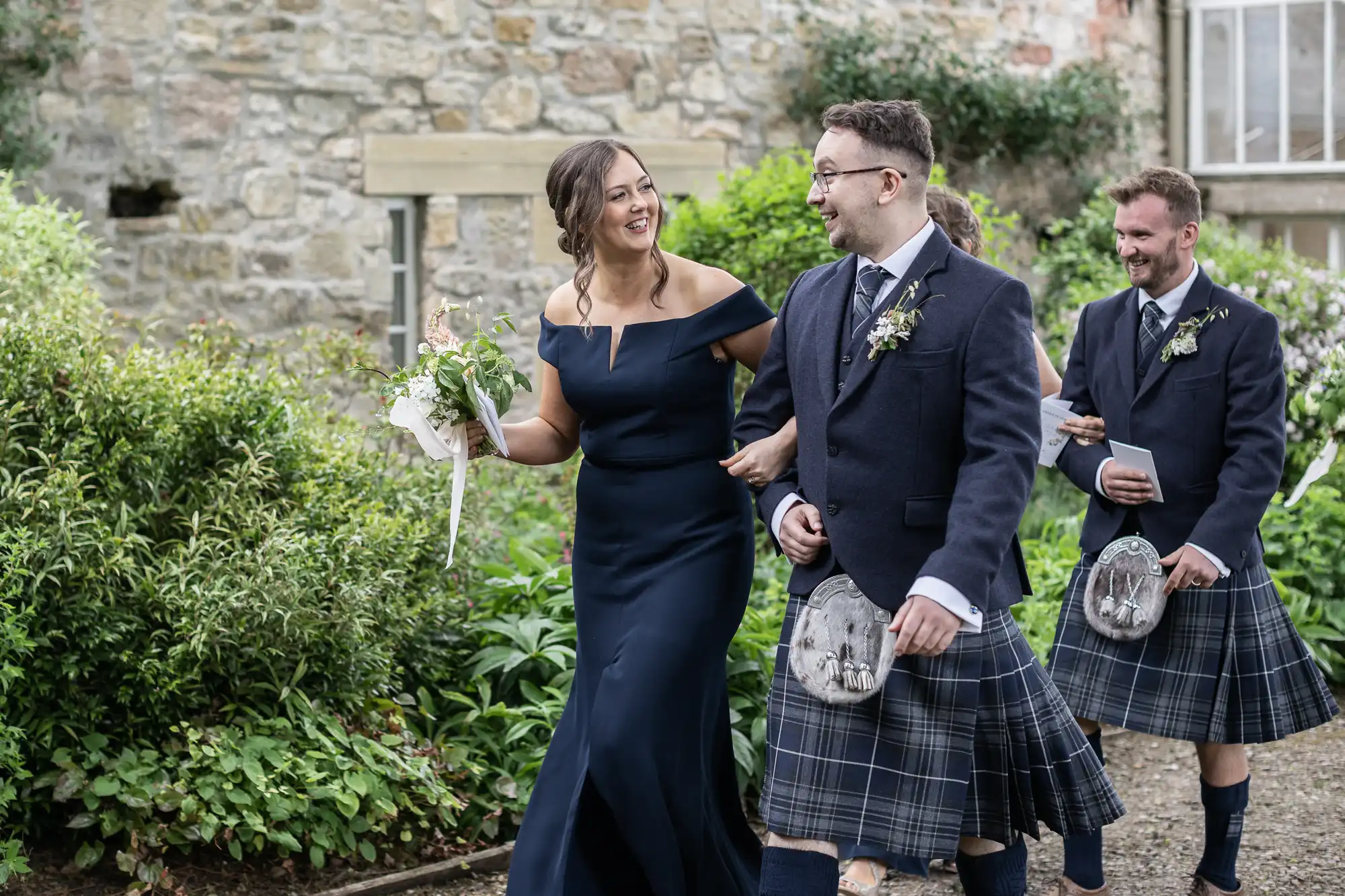 A bride in a blue dress holds hands with a groom in a kilt, laughing together with a man in a kilt and carrying a drink in the background, outside a stone building.