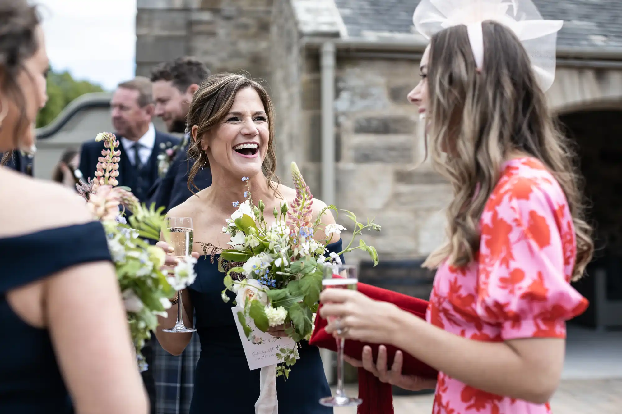 A joyous woman in a white dress holding a champagne glass and flower bouquet laughs with another woman in a red floral dress at a wedding.