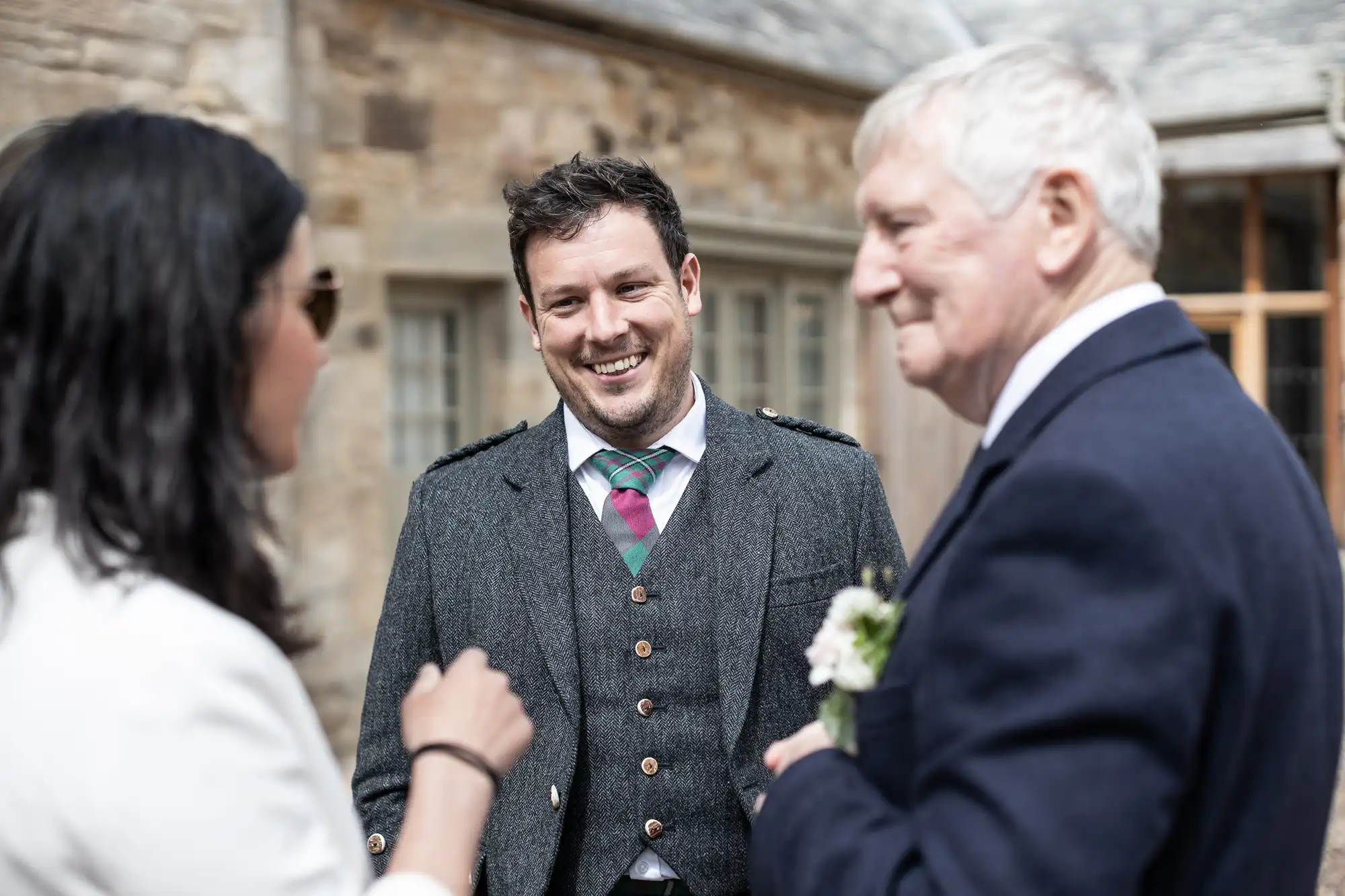 Three people conversing outdoors; a smiling man in a tweed suit with a boutonniere stands between an older man and a woman partially visible.