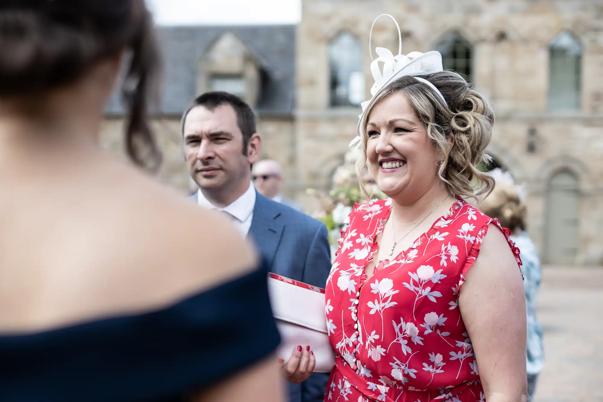 A smiling woman in a red floral dress and white fascinator at an outdoor wedding ceremony, with others around her.