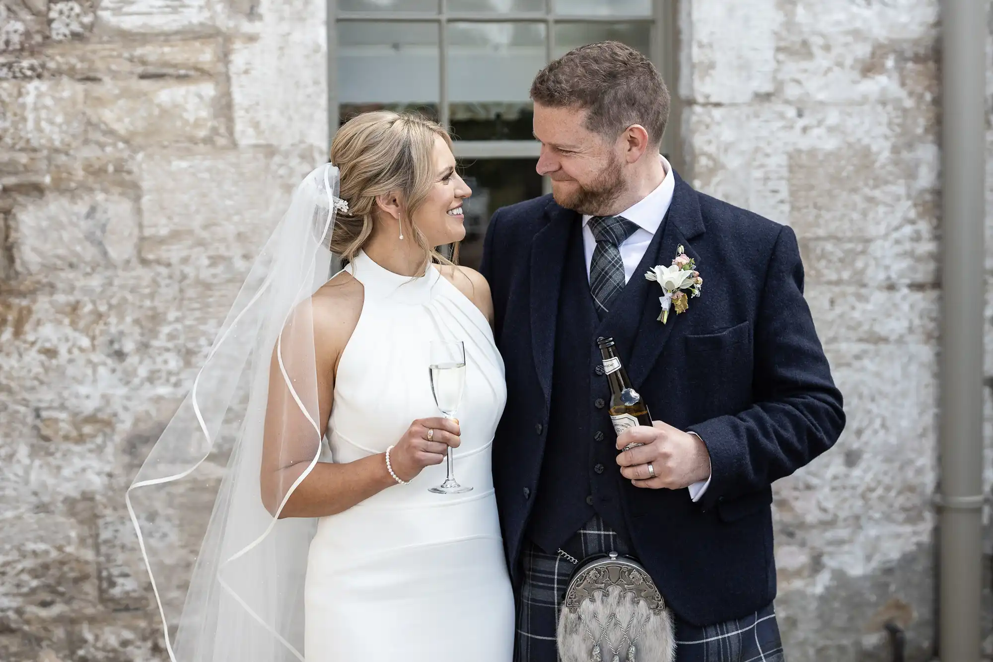 Bride in a white dress and groom in a kilt smiling at each other while holding champagne glasses at a wedding ceremony.