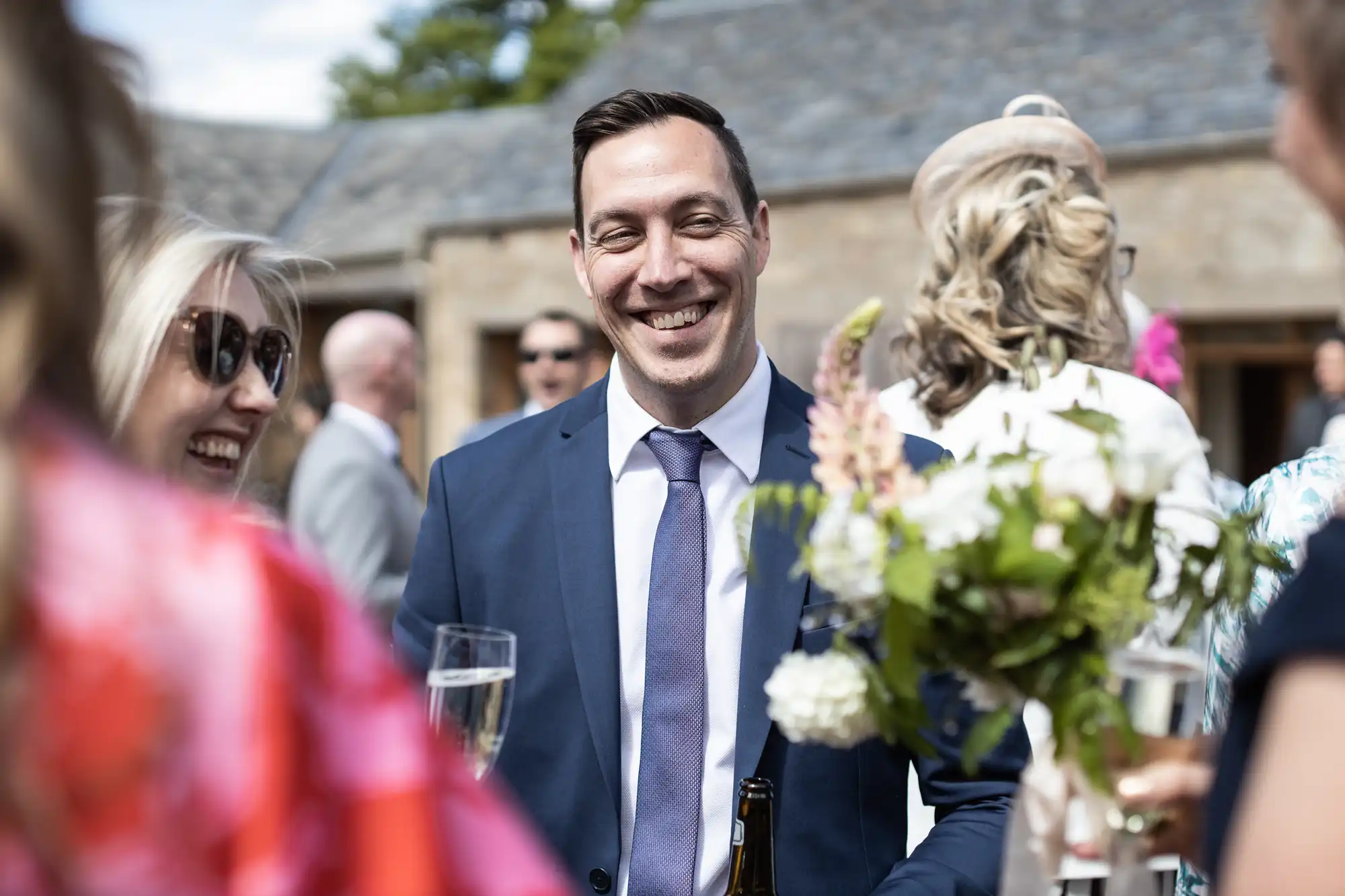 A man in a suit smiling at a wedding reception, surrounded by guests and floral decorations in a sunny outdoor setting.