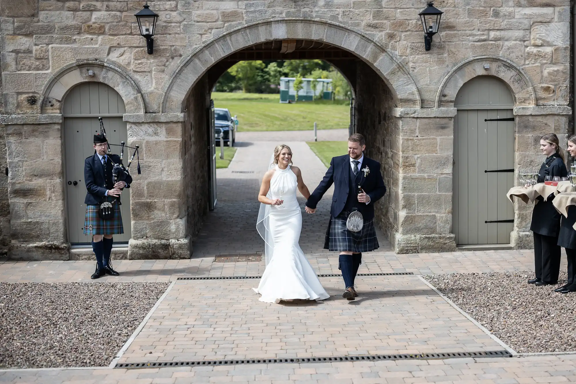 A bride and groom, holding hands and smiling, walk through an arched gateway, flanked by two people in traditional Scottish kilts.