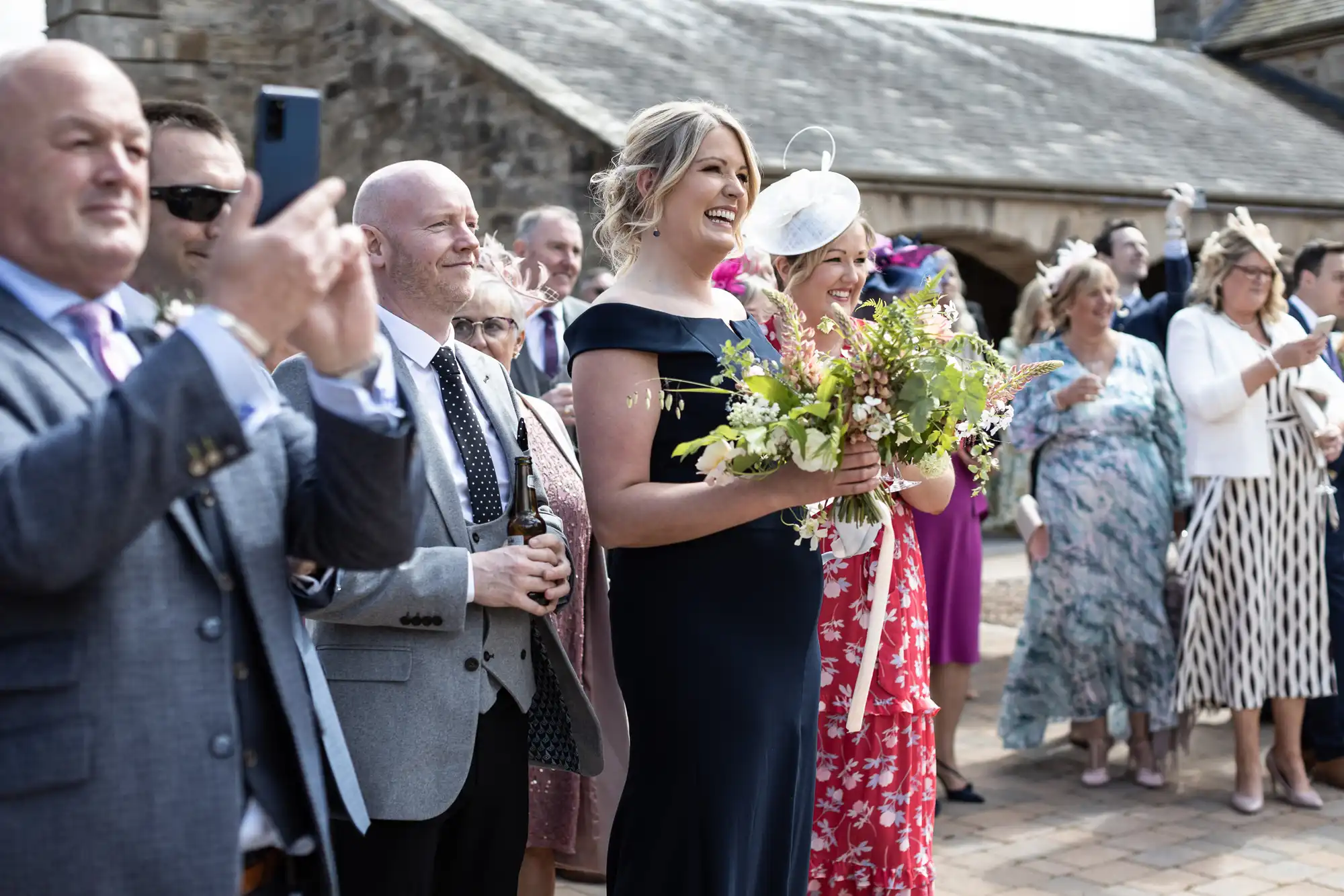 Wedding guests smiling and clapping as they watch a ceremony outside a stone building, some holding phones to capture the event.