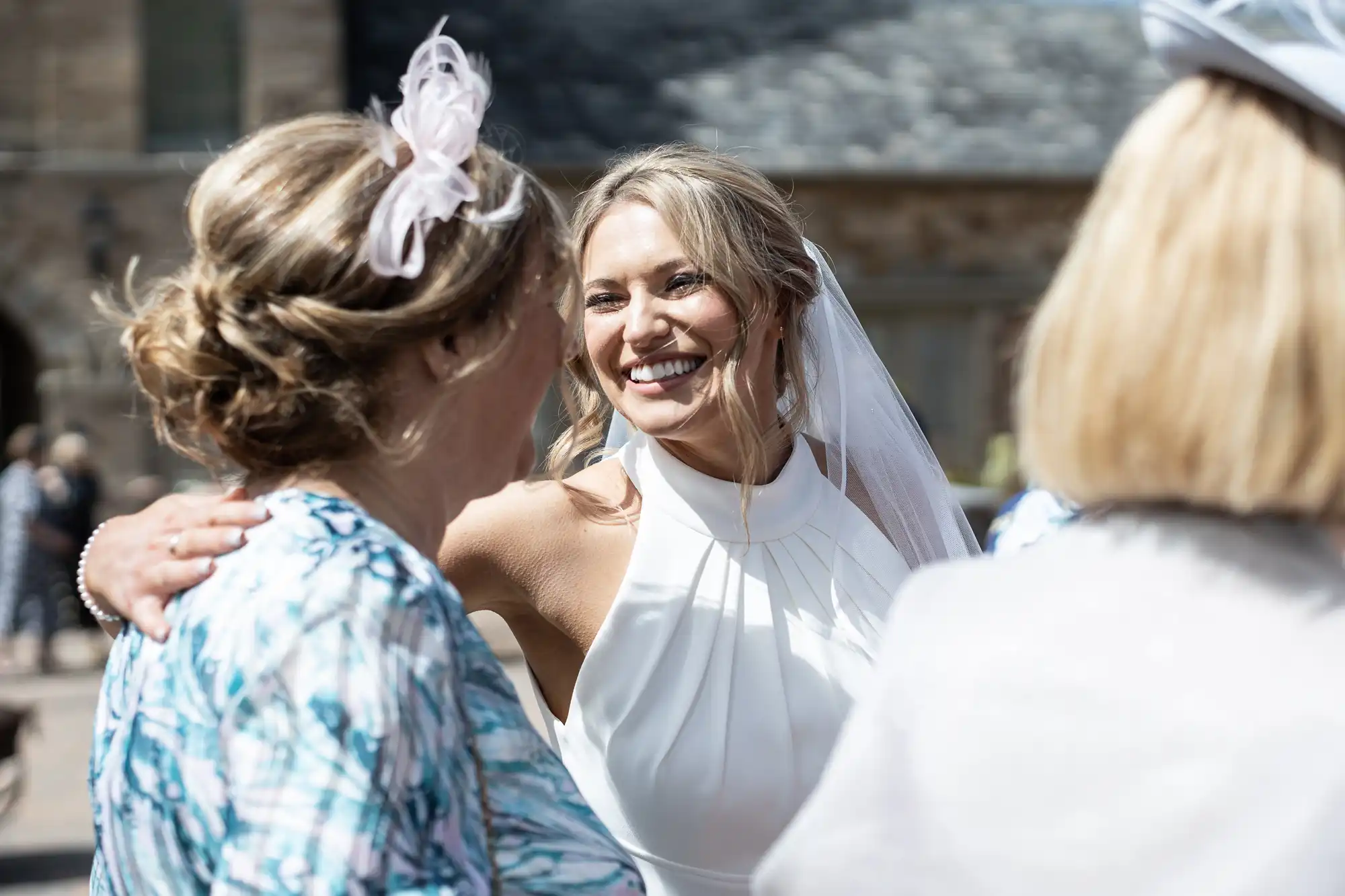 A bride in a white dress and veil smiles joyfully while talking with two women in formal attire at an outdoor event.