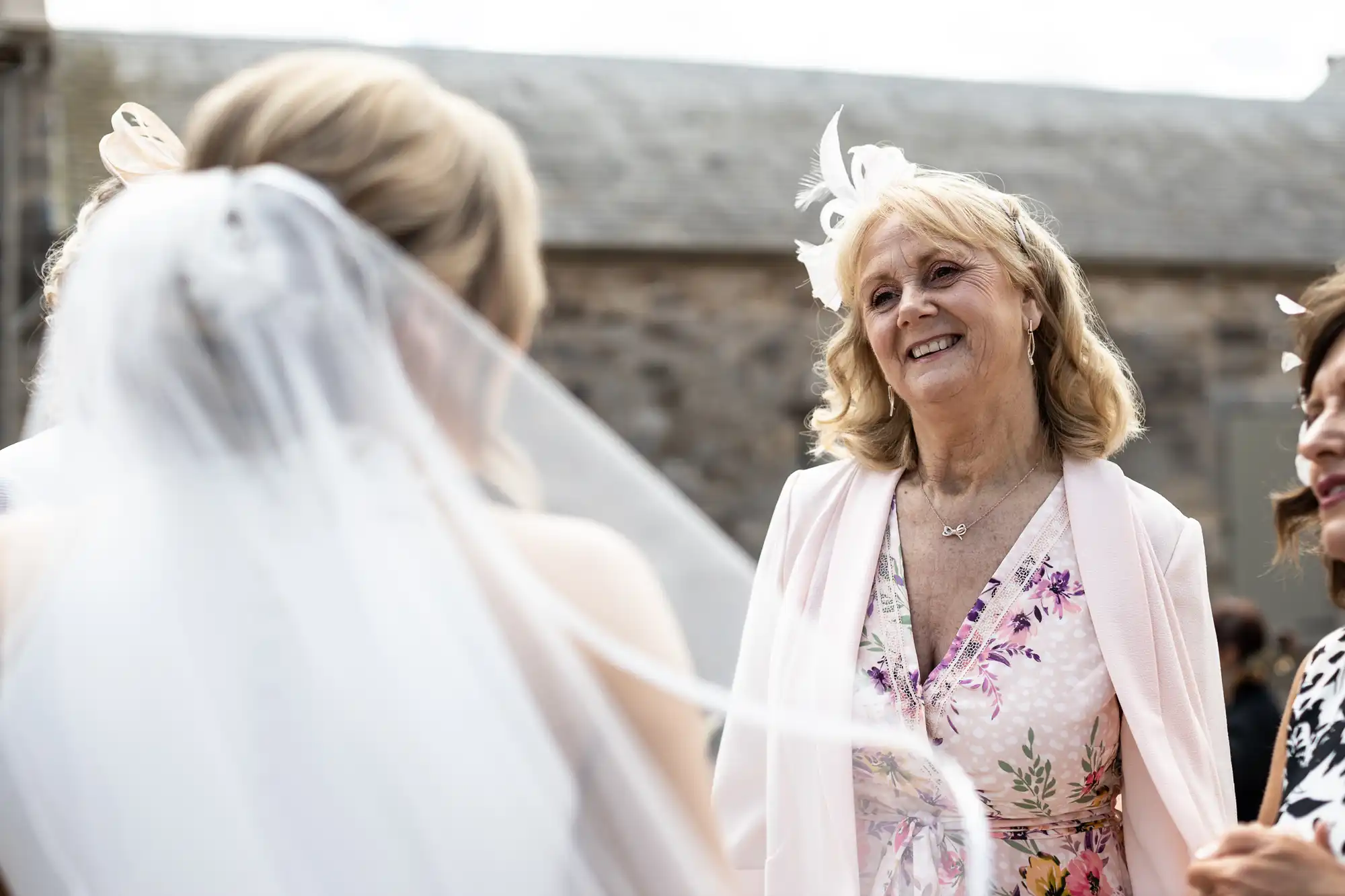 An older woman with blonde hair in a floral dress smiles at a bride wearing a veil, at an outdoor wedding ceremony.