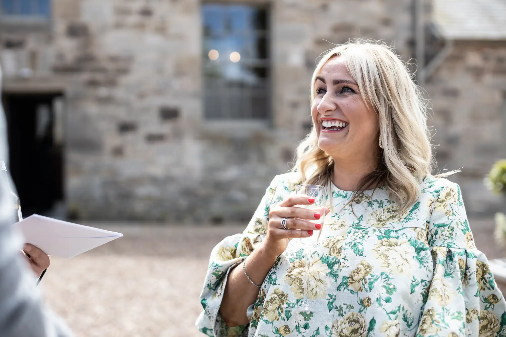 A woman in a floral dress laughs while holding a glass of wine at an outdoor event, with a historic stone building in the background.