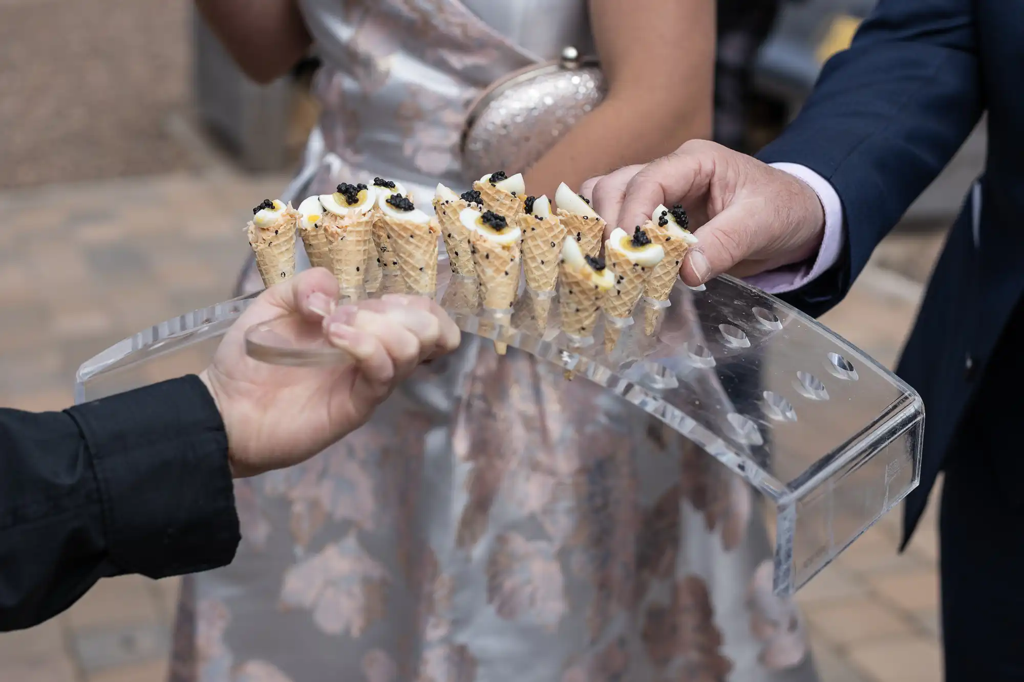 Guests at a wedding reception taking caviar appetizers served on clear glass tray, with focus on hands and food.