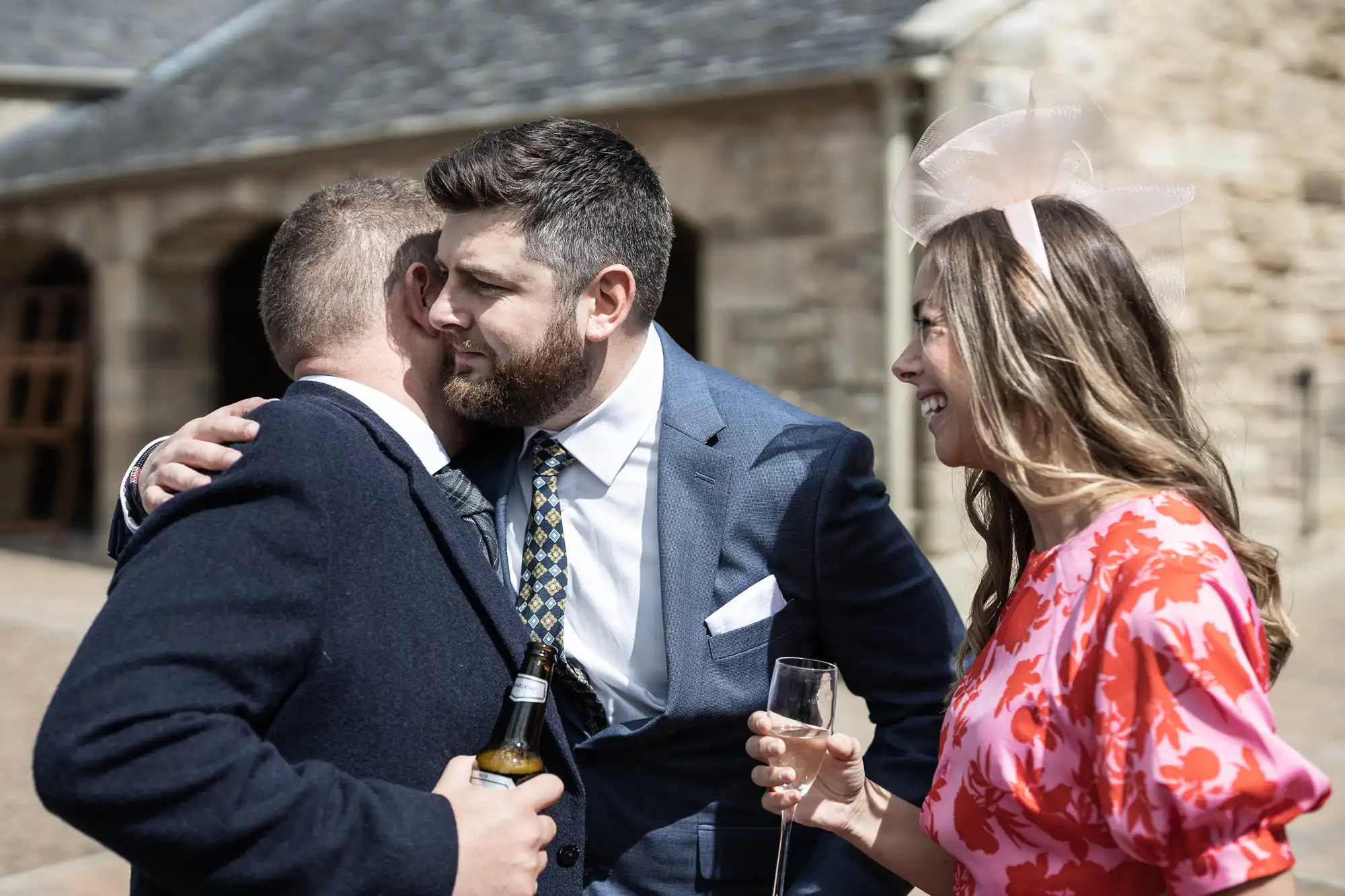 A man in a suit embraces another man while a smiling woman in a floral dress watches, holding a champagne flute. They are at an outdoor event with historic stone buildings in the background.