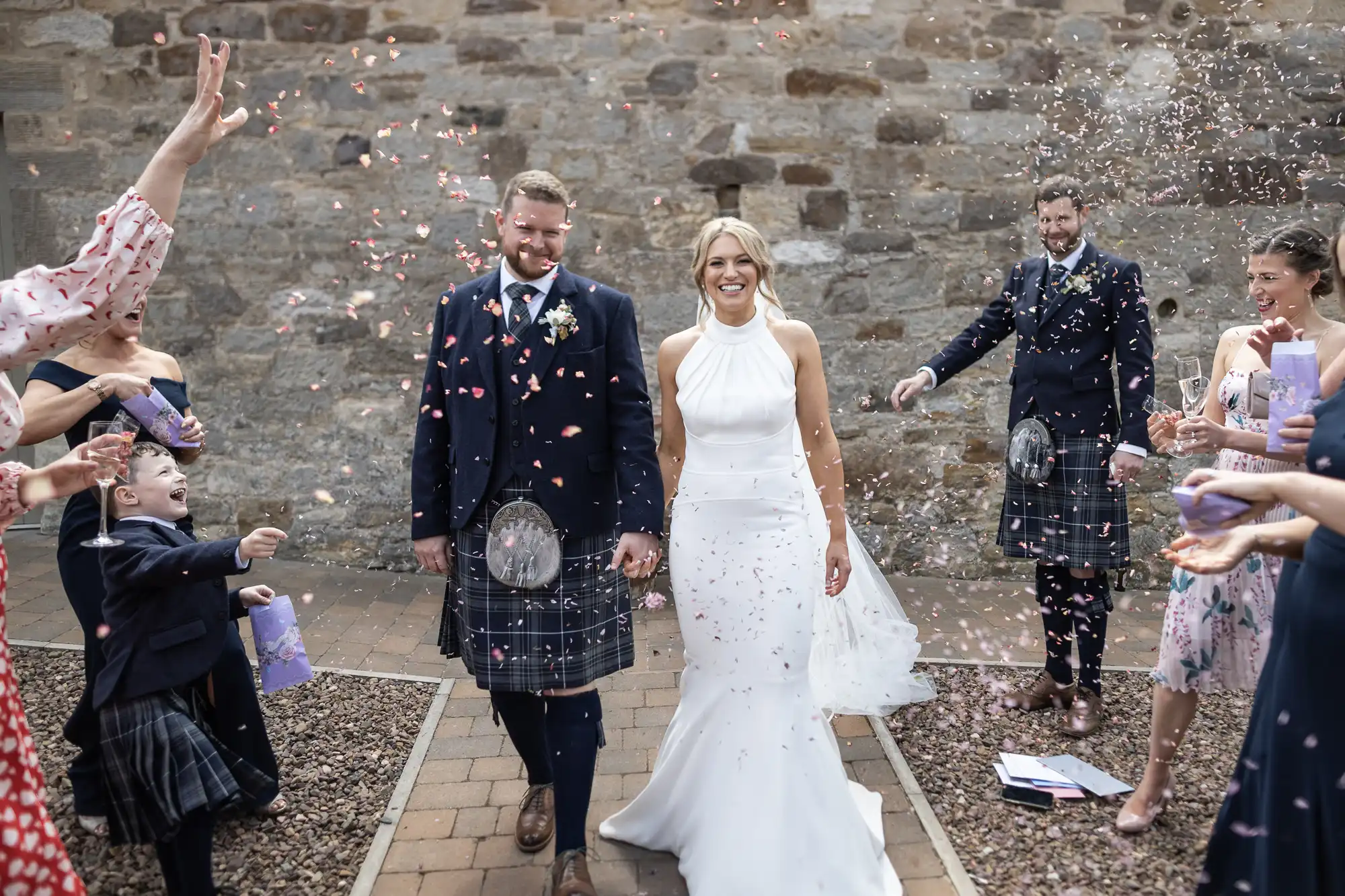 A newlywed couple walks through a shower of confetti, smiling, surrounded by guests and a child joyfully throwing confetti in an old stone courtyard.