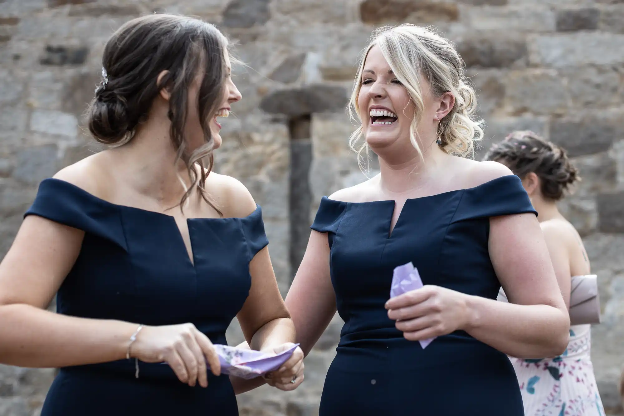 Two women in navy blue dresses laughing joyfully at an outdoor event, holding small purple papers.