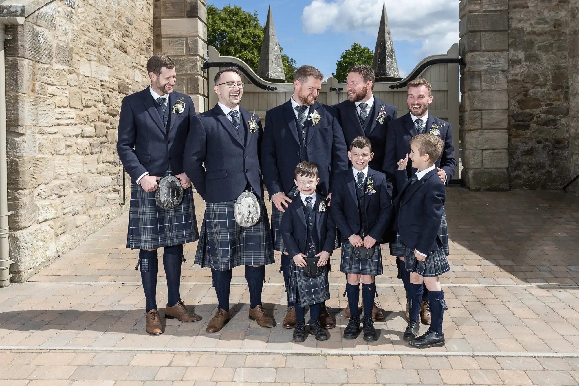 Five men and three boys in formal Scottish attire, including kilts and sporran, posing happily outside a stone building.