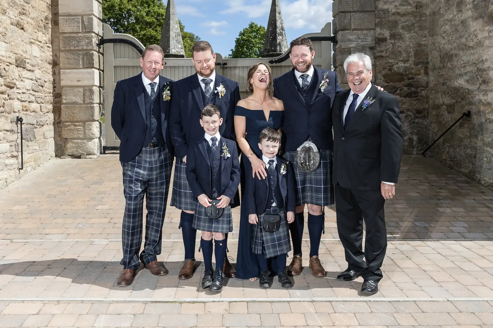 Group of six men and one woman in formal attire posing joyfully outside, with three children in front; all are wearing tartan kilts.