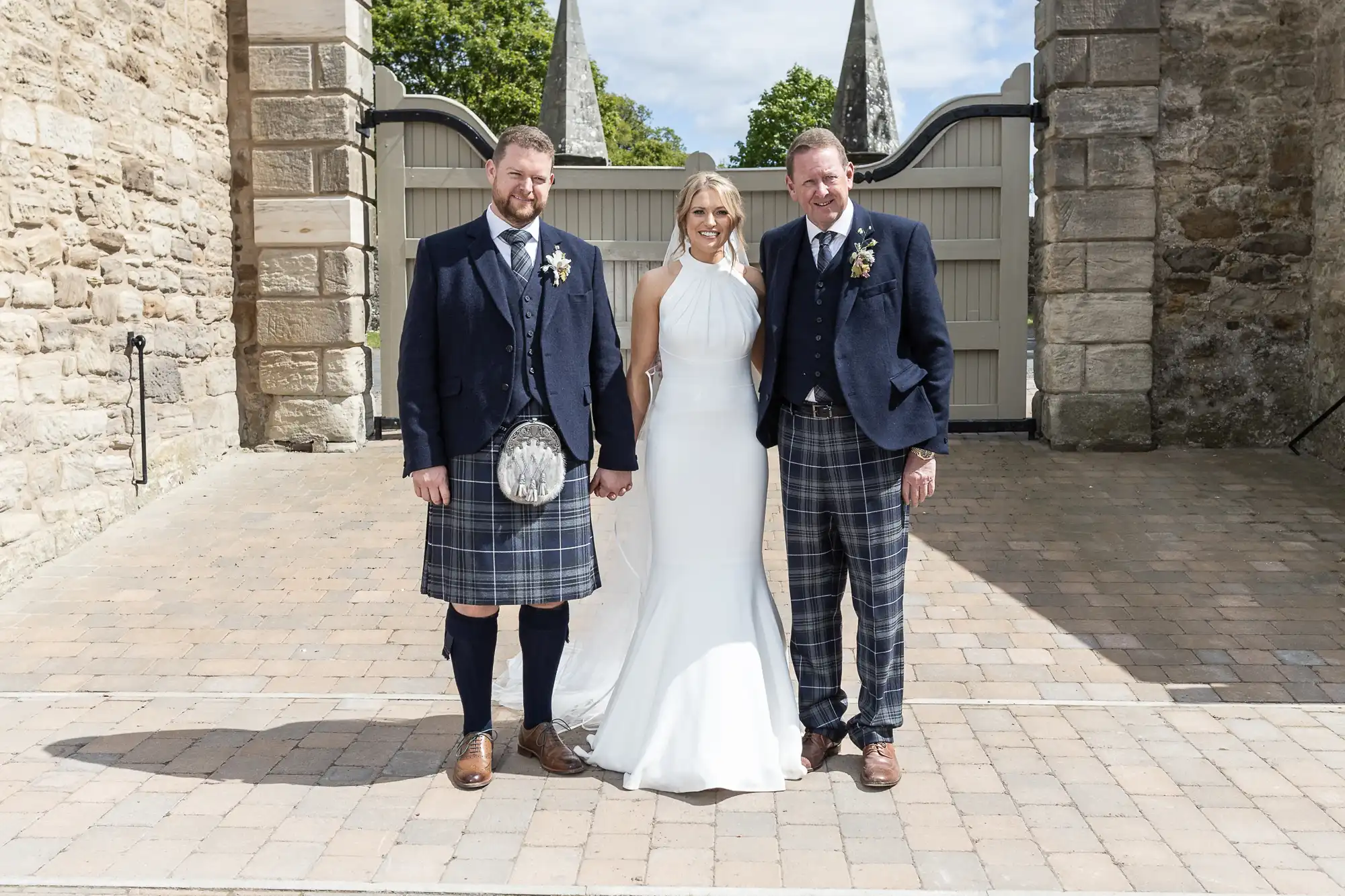 A bride in a white dress flanked by two men in traditional Scottish kilts and jackets, standing outside a stone building entrance.