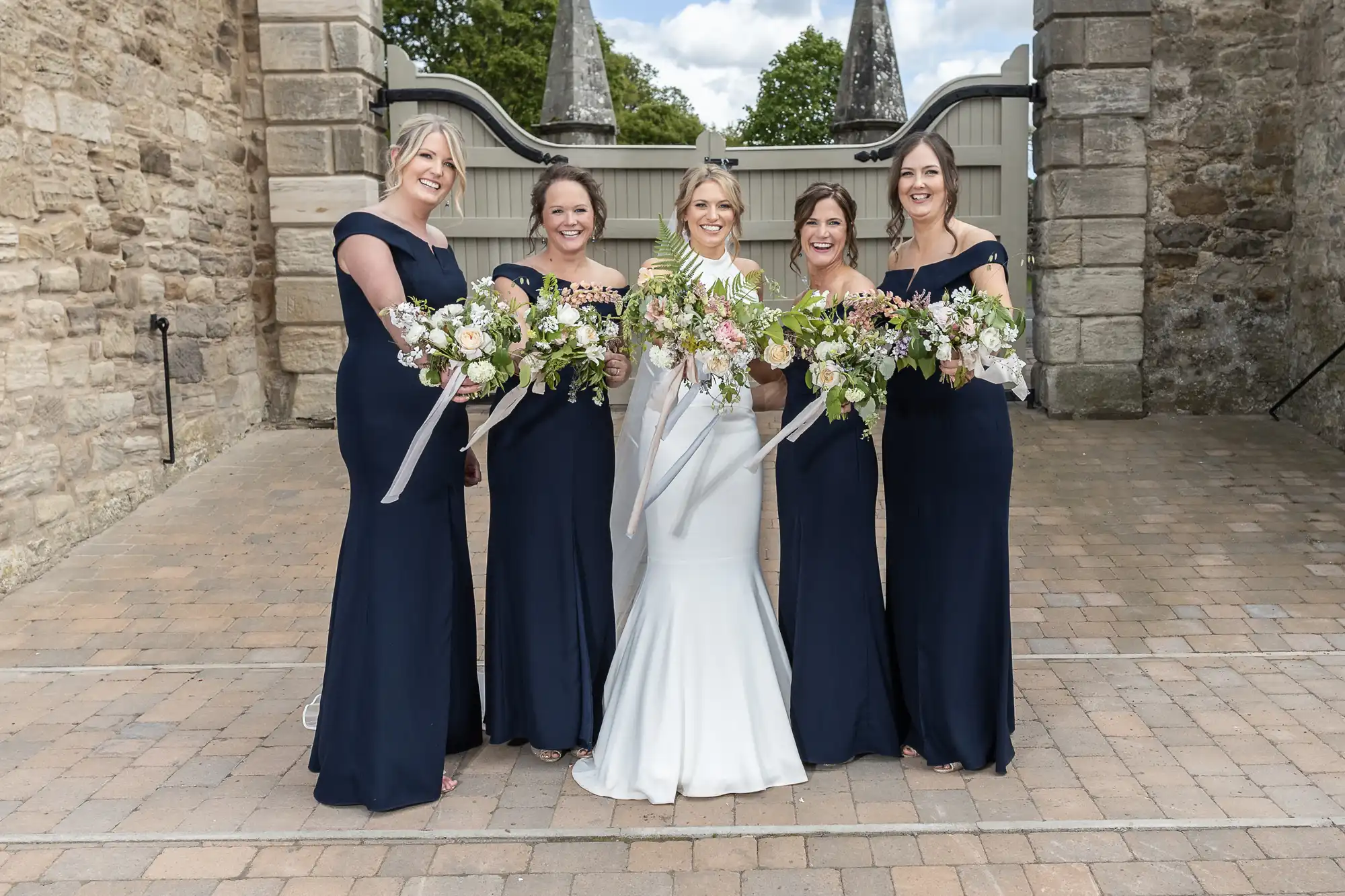 Five women in formal wear posing with bouquets at a wedding, with the bride in a white dress, flanked by four bridesmaids in navy blue dresses.