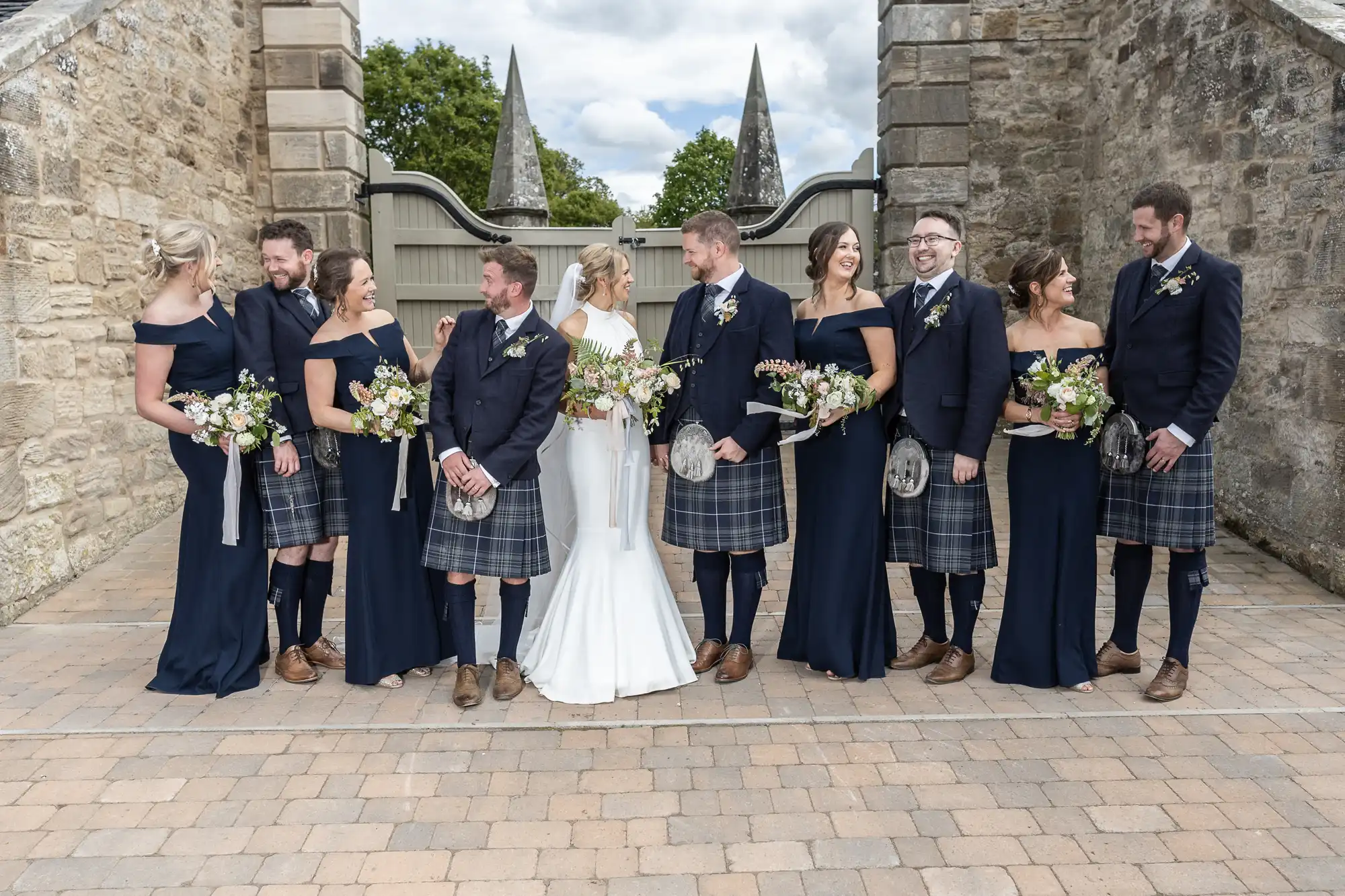 A wedding party consisting of six men in kilts and five women in navy dresses smiling and posing outdoors with bouquets.