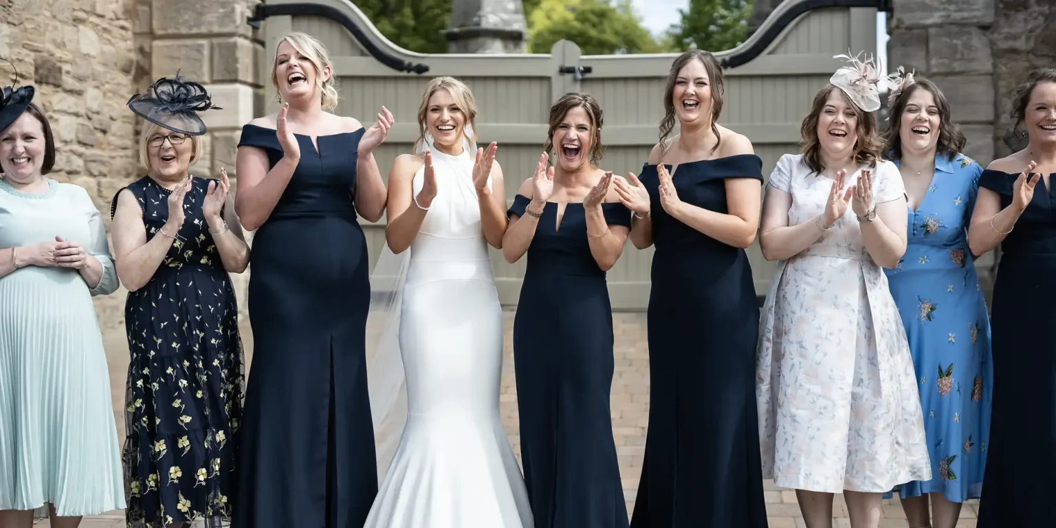 A group of women in elegant dresses, including one in a white gown, cheerfully clapping at a wedding ceremony.