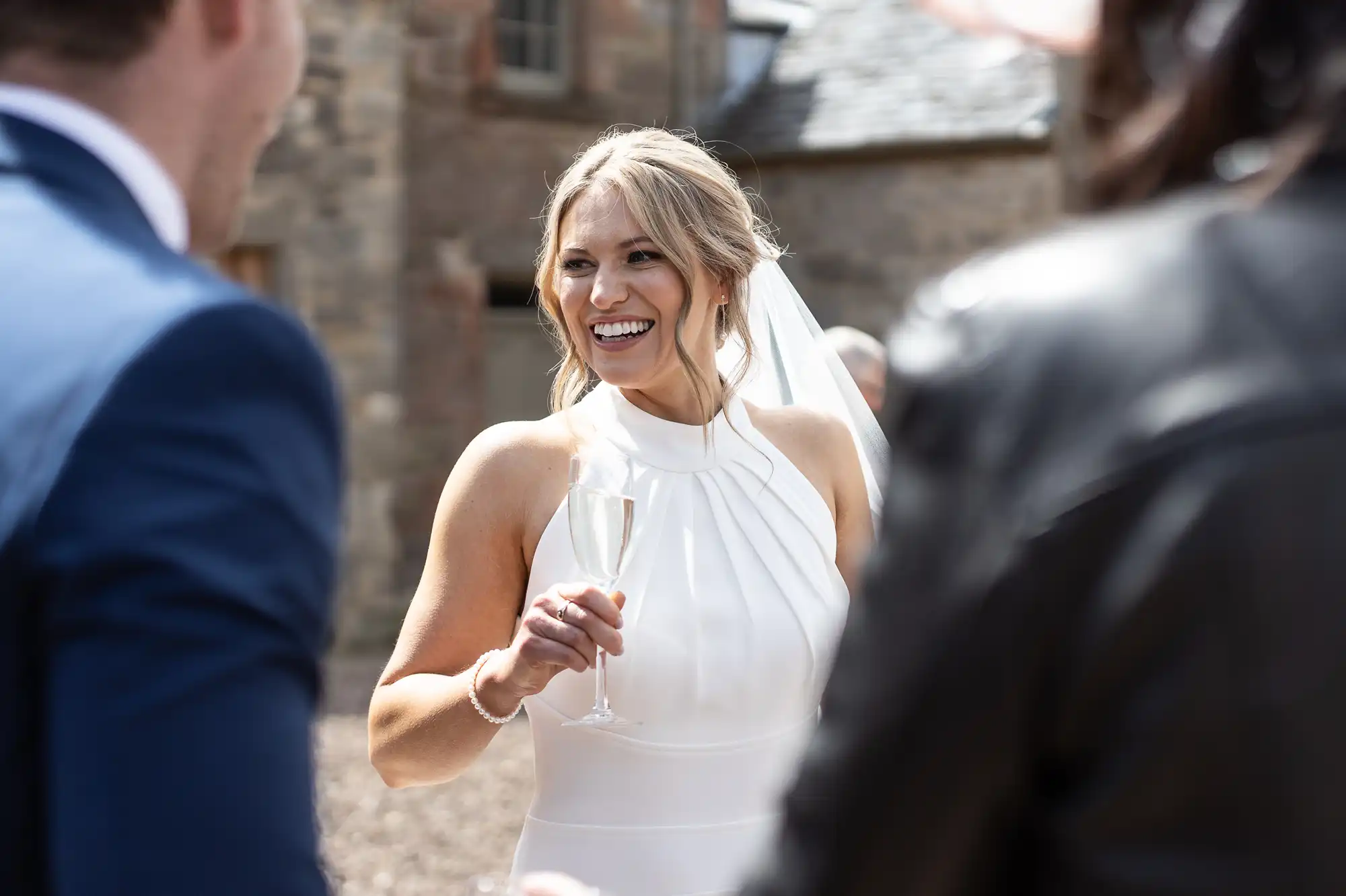 A joyful bride holding a champagne flute, laughing during a conversation with guests at an outdoor wedding setting.
