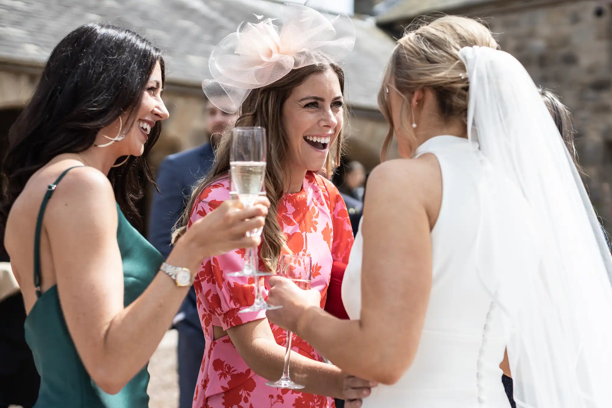 Three women laughing and conversing at a wedding; two guests in dresses holding champagne glasses and the bride in a white gown.